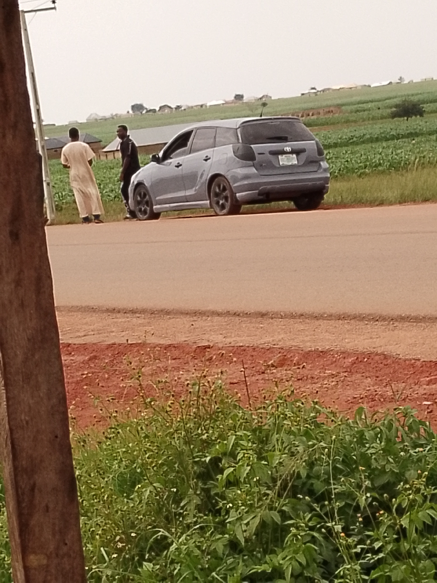 Two men are standing next to a gray car on the side of a road. The man on the left is wearing a traditional light-colored robe, and the man on the right is wearing dark clothing with a white stripe on the sleeves. The car is a hatchback with tinted windows and black rims. Behind the car, there is a green field of crops, and in the distance, there are some buildings. The sky is overcast, suggesting it might be a cloudy day. The foreground shows a wooden pole and some green vegetation. The scene appears to be in a rural or semi-rural area. No specific activity or event is clearly taking place, and the individuals' emotions are not discernible. The license plate on the car is partially visible but not legible. The location is identified as Agwala, Nigeria.