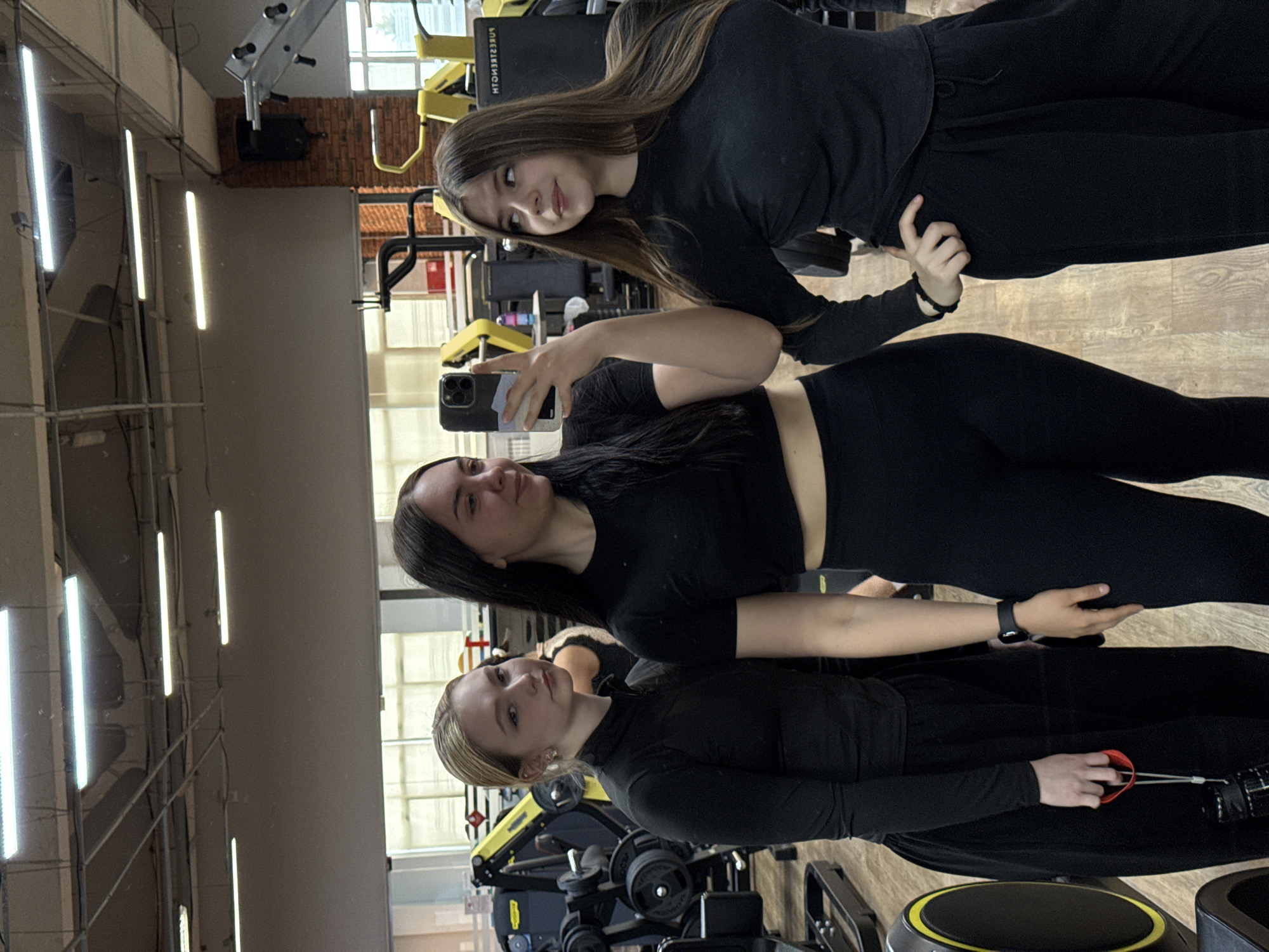 Three women are in a gym in the city of Chaikovsky, Russia. They are posing for a selfie. The woman in the middle is holding the phone. They are all dressed in black workout clothes. Behind them are various weight machines and dumbbells. The gym has bright overhead lights.