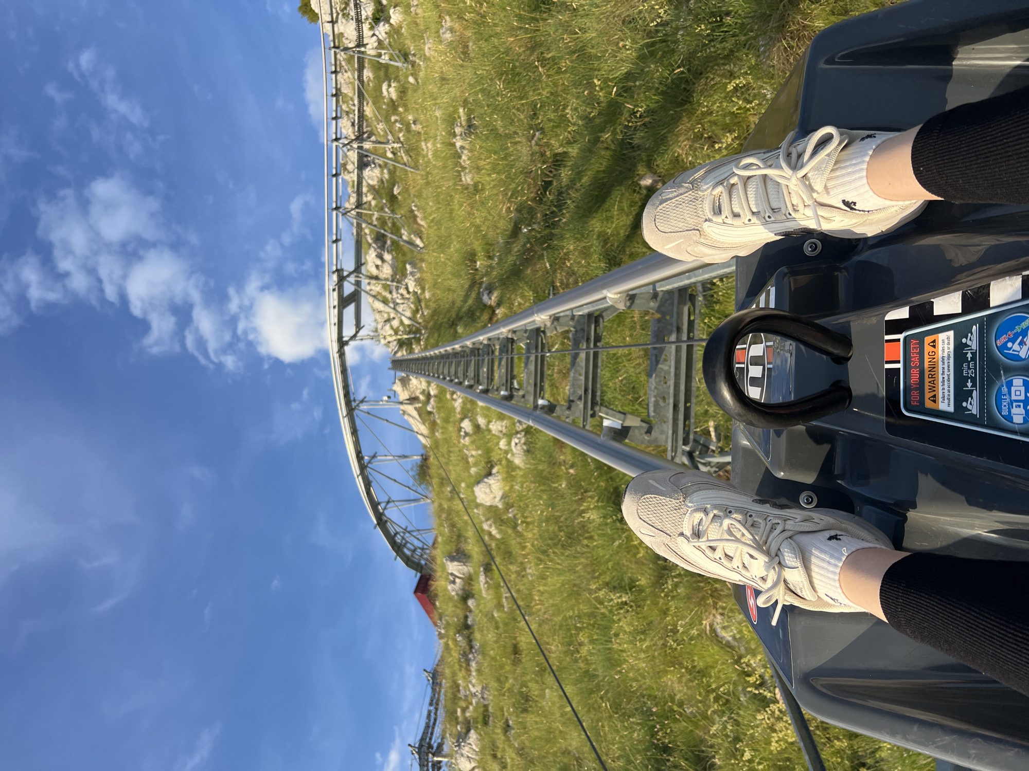 This first-person perspective image captures the ascent of an alpine coaster on a sunny day in Kuk, Montenegro. The viewer's feet, clad in off-white athletic shoes and white socks with a small dark emblem, are positioned on the dark grey floor of the coaster cart. The cart is actively climbing a sturdy metal track, which extends steeply uphill and curves to the right in the distance. The track is supported by an open metal framework.
The surrounding environment consists of a verdant hillside covered in lush green grass, dotted with numerous light-colored rocks and boulders. In the mid-ground to the left, a small structure with a distinct red roof is visible amidst the rocky, grassy terrain. The sky above is clear and bright blue, featuring scattered white cumulus clouds.
The front panel of the coaster cart displays several informational and safety decals. A prominent orange triangular warning sign reads "FOR YOUR SAFETY A WARNING," accompanied by text advising adherence to safety rules to prevent injury or death. Additional pictograms illustrate safety guidelines, including minimum height requirements (e.g., "min 25 m"), a speed limit (e.g., "25 mph"), and a "BUCKLE UP" instruction. A black handle with red detailing is visible on the right side of the cart's forward section. The rider's lower legs are clad in black ribbed fabric, likely trousers or leggings.