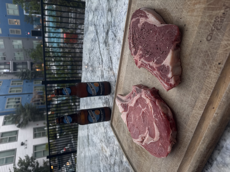 The image depicts an outdoor residential scene in Gainesville, United States of America, likely on a patio or balcony overlooking a communal area. In the foreground, a light-brown wooden cutting board, visibly worn with knife marks and bearing the "Chicago Cutlery" brand on its lower right, rests on a mottled light-colored stone countertop. On the cutting board are two raw beef steaks: one bone-in ribeye on the left, and a boneless cut on the right, which is generously seasoned with coarse black pepper.
Behind the cutting board, two open amber glass bottles of "Blue Moon Belgian White" beer, each with an orange slice depicted on its label, stand upright on the same stone countertop. Further back, a black metal fence separates the immediate area from a light-blue swimming pool. In the background, multiple multi-story residential buildings are visible; one section is white or light grey, and another prominent section is light blue, both featuring numerous windows and balconies. Green trees and foliage are interspersed throughout the background. The sky appears overcast or it is late in the day. No people are visible.