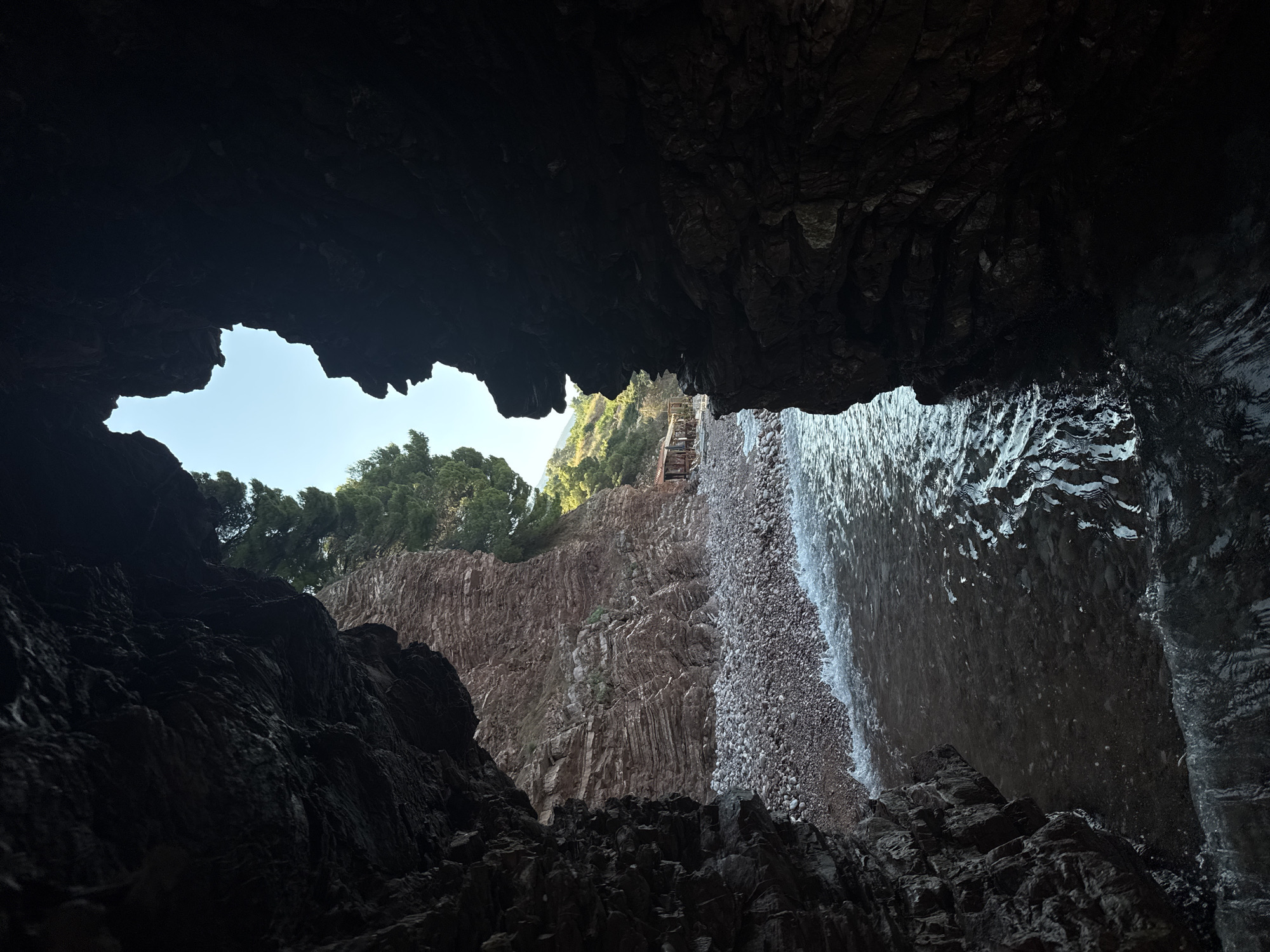 The image depicts a view from within a dark cave opening onto a pebble beach and the sea at Sveti Stefan, Montenegro. The foreground is dominated by the rough, dark rock walls of the cave, creating a natural frame for the scene beyond. The sea is calm, with small waves lapping at the shore. The beach is covered in small, smooth pebbles. In the background, a steep, layered rock face rises, topped with green trees. Further in the distance, along the edge of the beach, a row of rustic wooden structures, possibly beachside cafes or restaurants, are visible. The sky is clear and blue. The overall impression is one of natural beauty and coastal tranquility.
