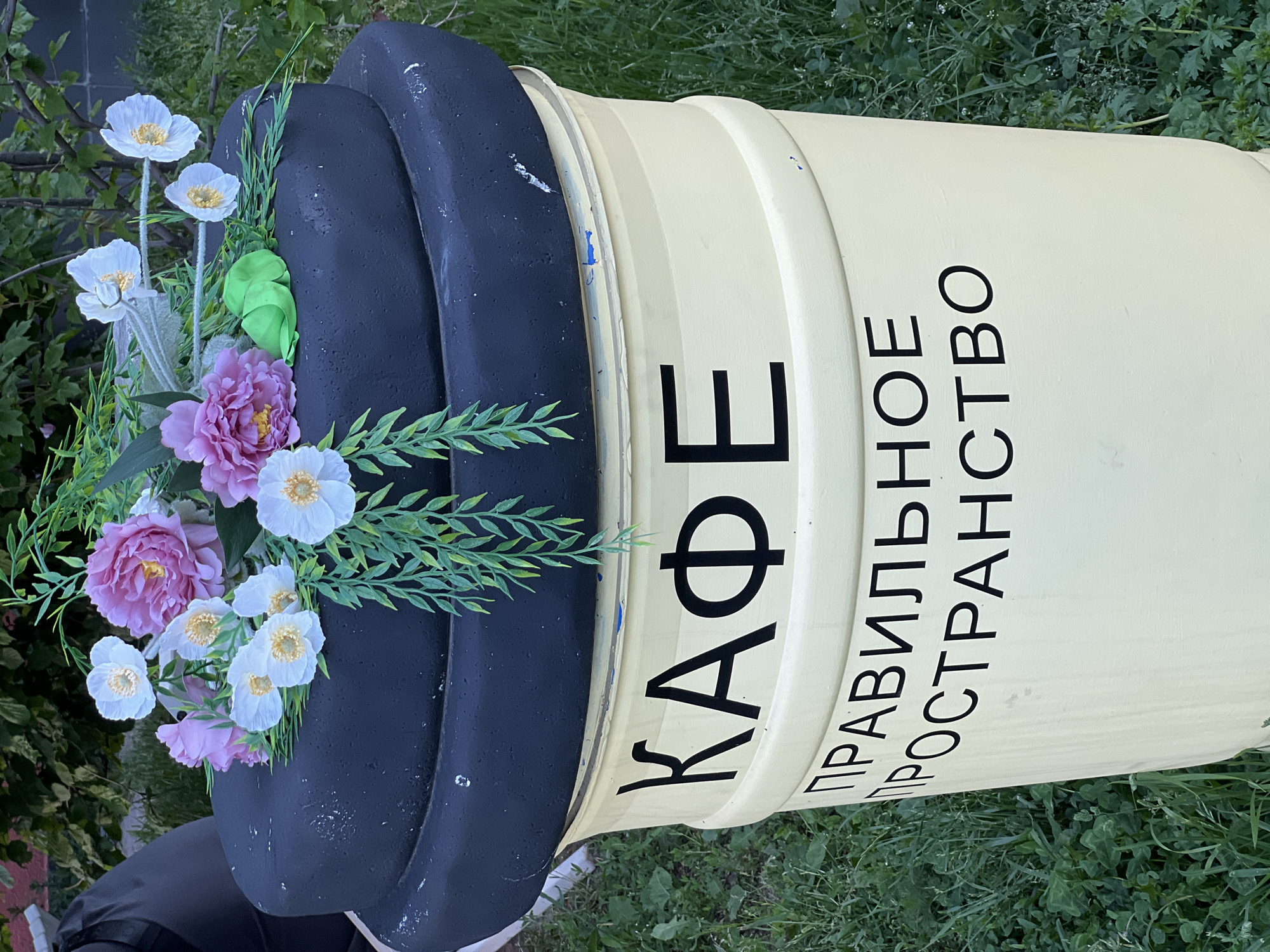 An outdoor, eye-level shot captures a large, decorative coffee cup sculpture lying horizontally on green grass, set in Mytishchi, Russia. The main body of the cup is a light yellow-cream color, featuring horizontal ridges. A black, curved element, resembling a lid or rim, is positioned on the left side of the cup.
Artificial flowers and green foliage are arranged within the black section of the sculpture, giving the impression of plants growing out of the cup. The flowers include several white blossoms with yellow centers and a few larger, pinkish-purple peony-like flowers. Green artificial leaves and grasses are interspersed among the blooms.
The background consists of blurred green foliage and grass, indicating a natural or landscaped setting. The lighting suggests daytime, possibly with overcast conditions or indirect sunlight, as there are no harsh shadows.
Visible text on the yellow-cream body of the cup is in black Cyrillic letters. It reads: "КАФЕ" (CAFE) and "ПРАВИЛЬНОЕ ПРОСТРАНСТВО" (RIGHT SPACE or CORRECT SPACE). The sculpture shows some signs of wear, with small blue and white scuff marks on its surface. There are no people visible, and the scene depicts a static decorative display.