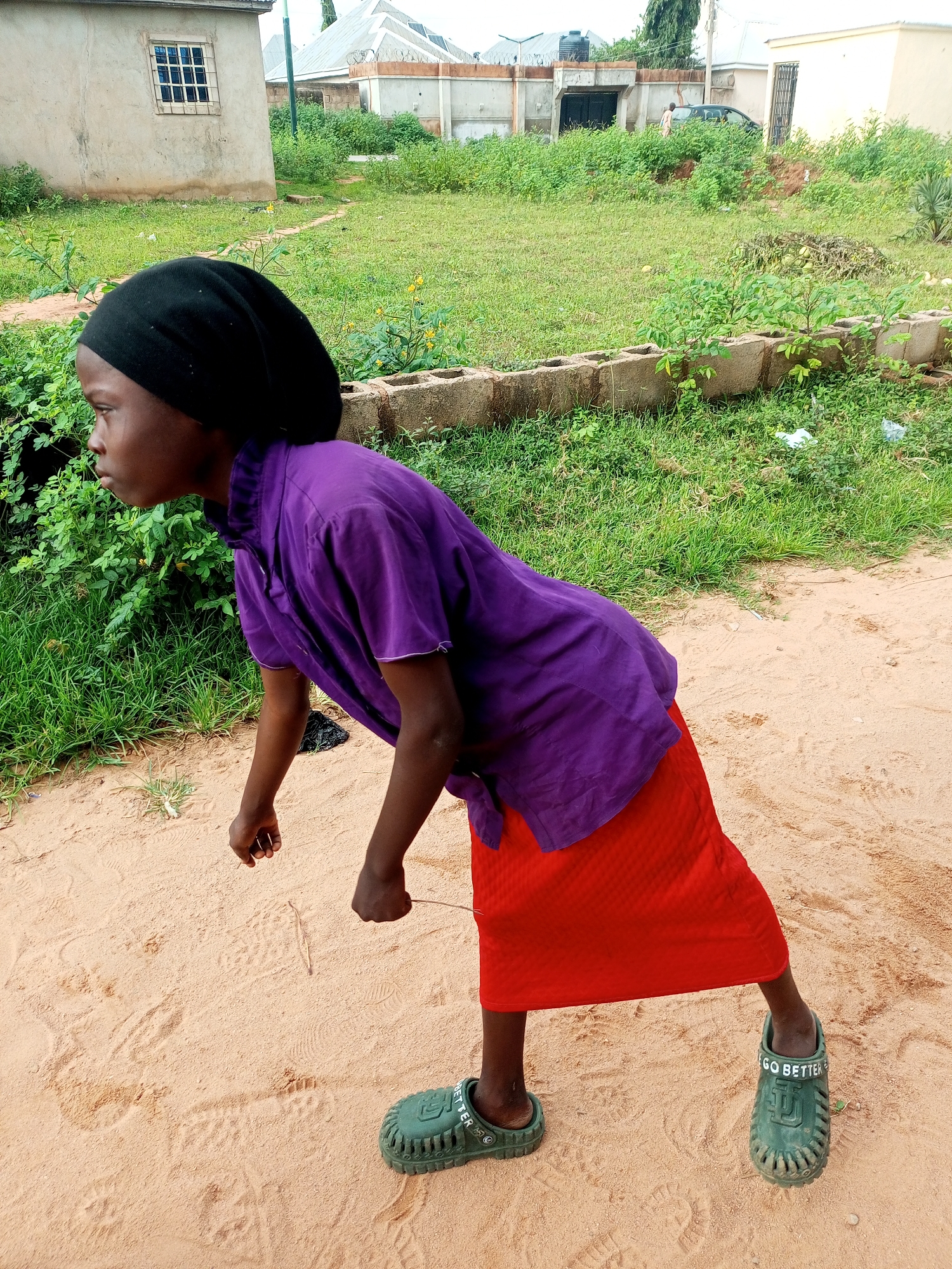 A young person wearing a black headscarf, a purple shirt, and a red skirt stands on a sandy dirt path. The person is wearing green clogs with the words "GO BETTER" printed on them. The background shows a grassy area with some buildings and trees. The scene appears to be outdoors in a semi-urban or rural setting.