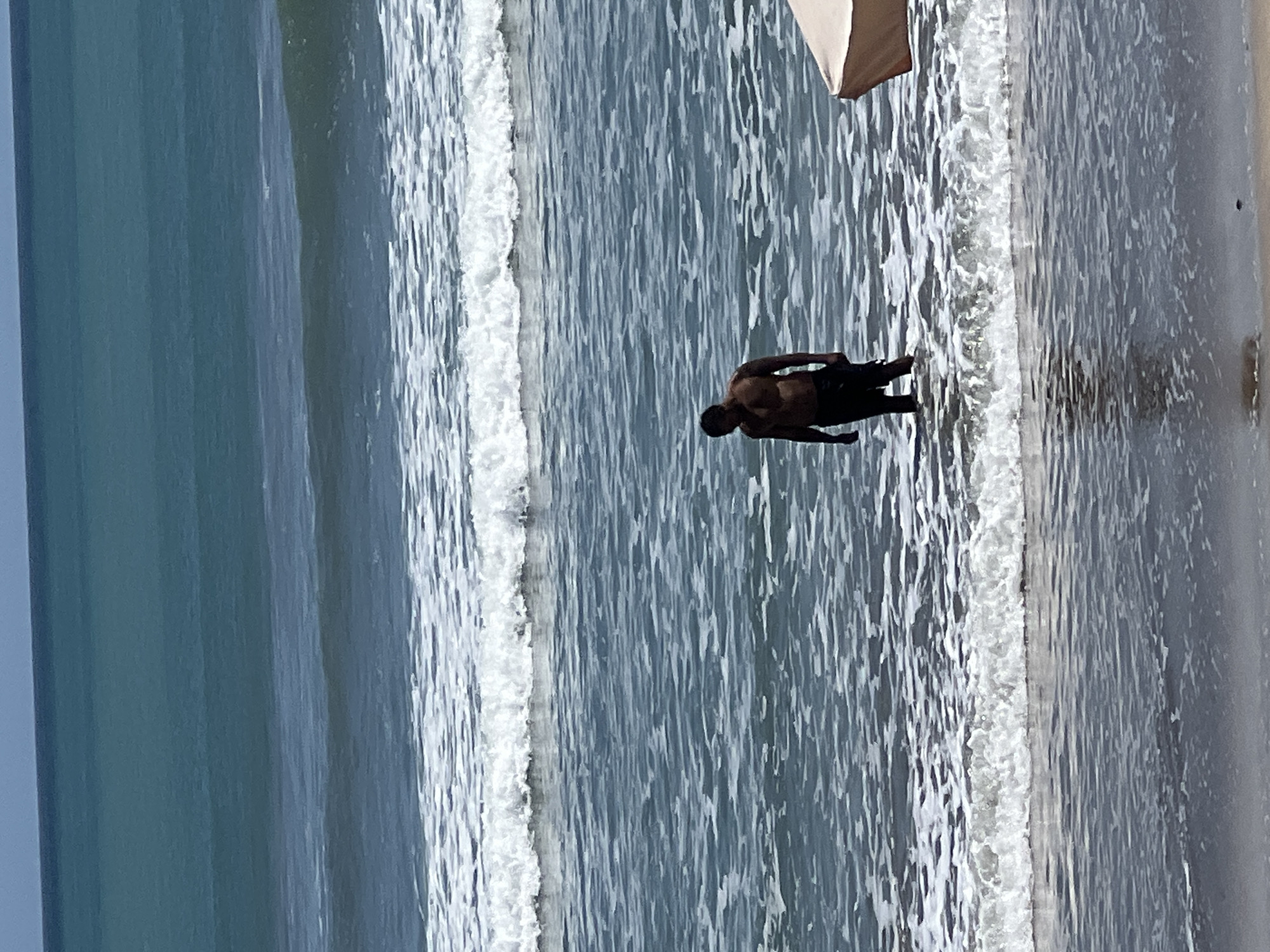 In Kuta, Indonesia, a lone male stands in the shallow ocean water. He is shirtless and wearing dark shorts. The waves are breaking around his legs, and the water appears to be a deep blue-green color. The sky is a clear, pale blue. In the lower right corner of the image, the edge of a large, light-colored umbrella is visible. The overall scene depicts a tranquil day at the beach with the ocean as the dominant element.