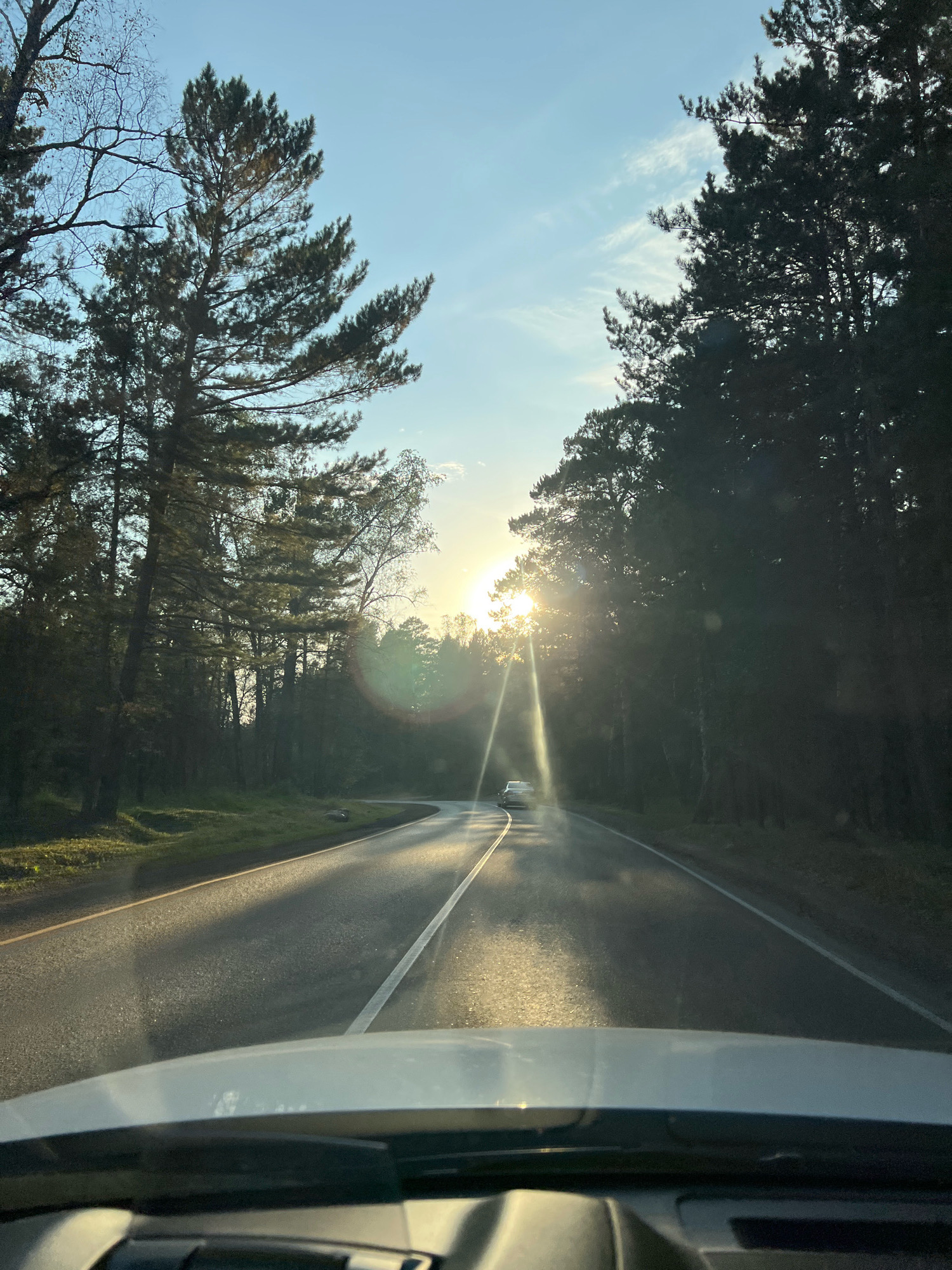 The image depicts a road curving through a forest on a sunny day, with the sun setting behind the trees. The view is from inside a vehicle, looking out through the windshield. The road is paved and marked with white lines. Several cars are visible on the road. The trees on either side of the road are tall and dense. The sky is blue with some clouds. The sun is low on the horizon, casting long shadows and creating lens flare. The overall impression is one of a scenic drive through a natural landscape.
This scene is likely located on a road outside of the city of Krasnoyarsk, Russia, possibly along the Yenisei River or in the surrounding taiga forests. The specific location cannot be precisely determined from the image alone.