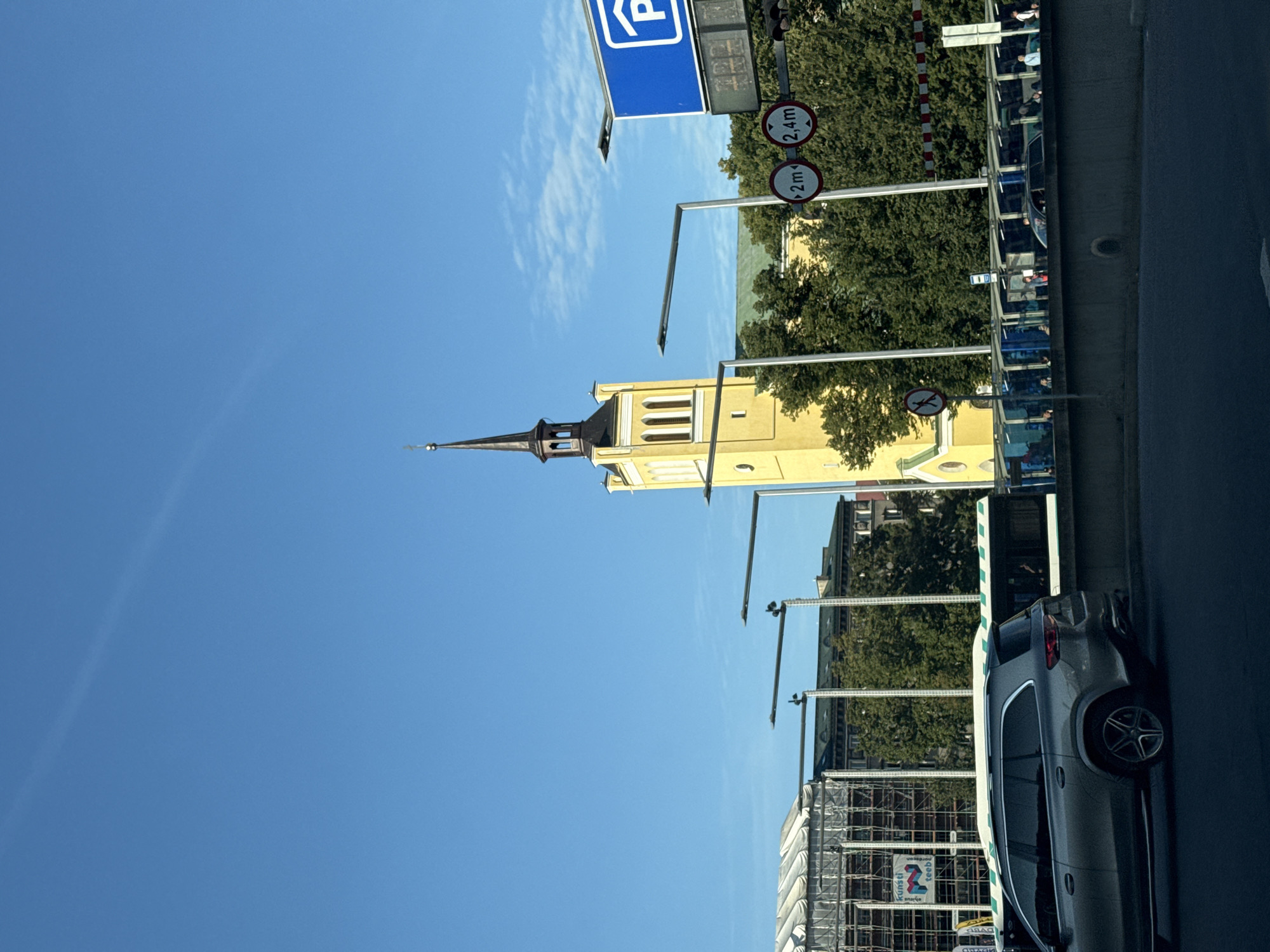 The image shows a street scene in Tallinn, Estonia, on a clear day. In the background, a tall, yellow church tower with a dark spire stands against a bright blue sky. To the left of the tower, a modern building, possibly under construction, is covered in scaffolding and white sheeting. In the middle ground, a row of trees separates the background from a bus stop with a blue glass enclosure. Several people are visible waiting at the bus stop. In the foreground, a silver SUV is parked on a road. Traffic signs are visible, including a blue parking sign with a white "P" and two circular signs indicating height restrictions of 2 meters and 2.4 meters. A street lamp with multiple lights is mounted on a pole. The overall impression is of an urban environment with a mix of historical architecture and modern infrastructure.