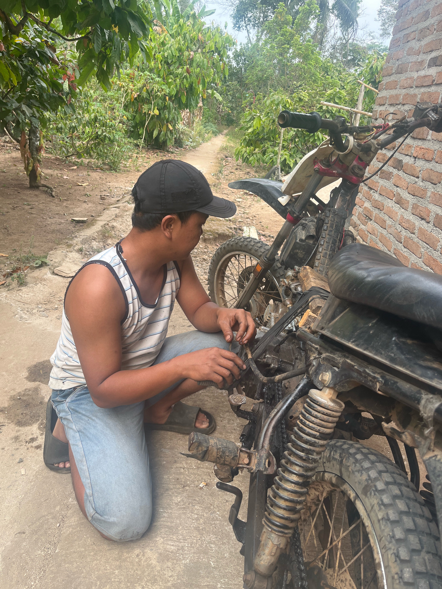 A man is kneeling next to a dirt bike, appearing to be working on it. He is wearing a black baseball cap, a white and blue striped tank top, and light blue denim shorts. He has dark skin and short black hair. The dirt bike is black and has some red accents. It is leaning against a brick wall. The ground is dirt and concrete. In the background, there are trees and foliage, suggesting a rural or semi-rural setting. A dirt path is visible leading away from the scene. The lighting suggests it is daytime. This scene is likely located in Kebumen, Indonesia, based on the context provided.