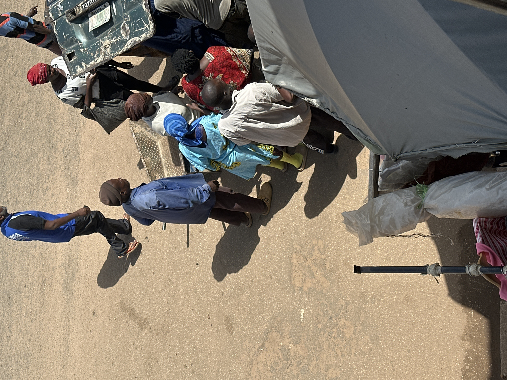 The image depicts an outdoor scene with several people gathered. In the foreground, a large gray tarp covers part of the frame, with a black metal pole extending upwards from its base. To the left of the tarp, there are some white plastic bags and a pink striped cloth.
In the middle ground, a group of people are interacting. One man, wearing a light-colored shirt and dark pants with white stripes, is bent over. He appears to be looking down at something or someone. Beside him, a woman in a red and white patterned dress is also facing downwards. Another woman in a bright blue headscarf and blue dress is looking towards the man. To their left, a man in a blue jacket and brown pants is looking away.
Further back, a man in a blue vest with "LOBA" visible on it walks away from the camera. Next to him, a woman in a white top with a red headscarf is holding a dark bag. There is also a green object that resembles a car part with "TOYOTA" written on it.
The ground is paved with a light-colored, textured material, likely asphalt or concrete, suggesting an urban or semi-urban environment. The lighting indicates it is daytime, with strong shadows cast by the people and objects, suggesting the sun is high in the sky. The overall impression is of a busy, possibly market or roadside gathering.
The location in Bukuru, Nigeria, suggests a West African context, which is consistent with the attire and appearance of the individuals in the photograph.