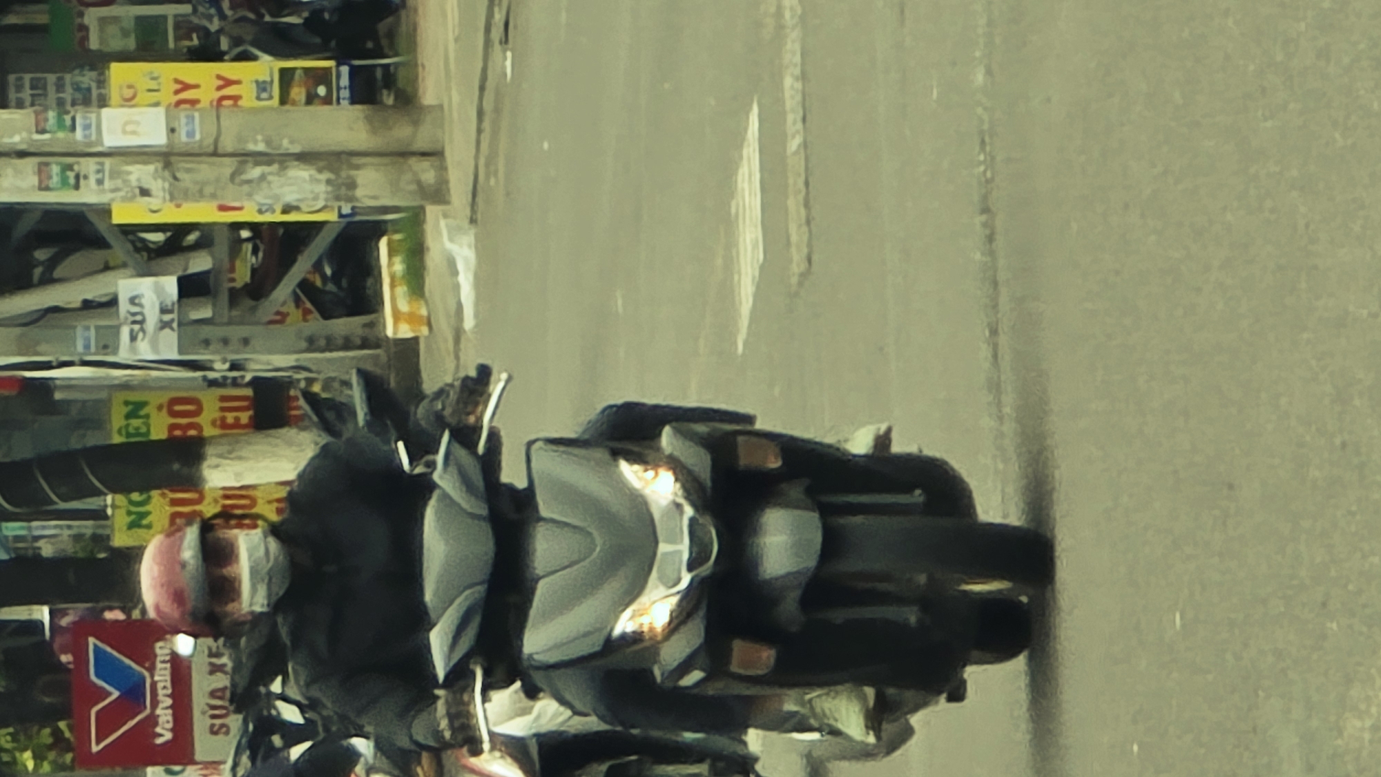 A person rides a dark-colored scooter on a paved road, captured during what appears to be daytime, with the scooter's headlights illuminated. The rider is wearing a reddish-pink helmet and a light-colored face mask. The scooter features grey and black body panels, with its front turn signals and main headlights visibly active.
To the left of the road, a bustling urban scene unfolds with numerous signs and banners affixed to buildings or walls. Several signs prominently display "SỬA XE," which translates from Vietnamese to "Repair Vehicle" or "Motorcycle Repair." A distinct red and white sign bears the "Valvoline" logo. Other partially visible Vietnamese text includes fragments like "EN BO EU," "QUY," and "BU BU." The background elements are somewhat blurry, suggesting motion or a shallow depth of field. The overall setting is indicative of a typical street in Vietnam, aligning with the provided context of Bac Nha Trang Ward. The image exhibits a slightly desaturated color scheme and some digital noise.