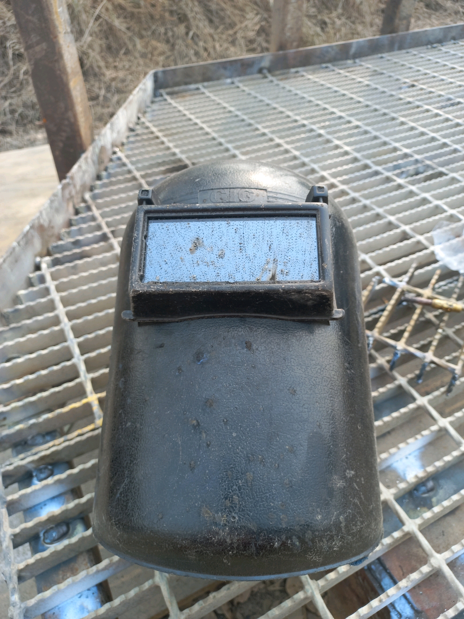 A black welding helmet rests on a metal grate. The helmet has a rectangular viewport with a blueish tint, speckled with dust. The grate is constructed of parallel metal bars with a diamond pattern in the gaps. In the background, dry, brown vegetation and a metal post are visible. The scene appears to be outdoors, with a muted, natural light. The location in Paringin, Indonesia is not discernible from the image.