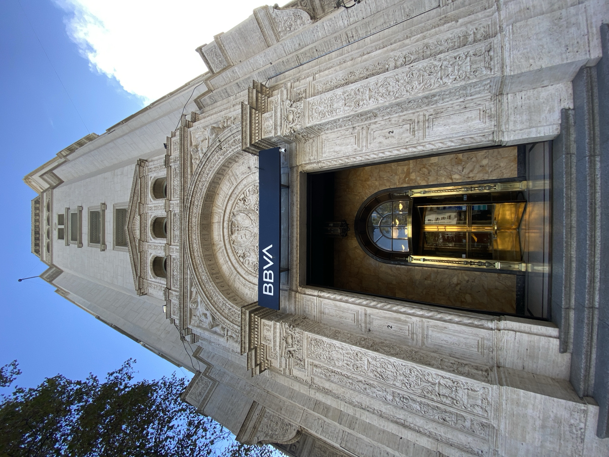 The image shows the entrance of a BBVA bank branch located in Buenos Aires, Argentina. The camera is positioned looking up at the building's ornate facade. The building is made of light-colored stone and features detailed carvings and architectural elements such as an arched entrance. A dark blue BBVA sign hangs above the doorway. Inside the entrance, a reflective golden door can be seen. The sky is visible at the top left of the frame with a blue gradient and a few clouds. Trees are visible at the bottom left corner of the image. A number "2" is on a stone block near the entrance.