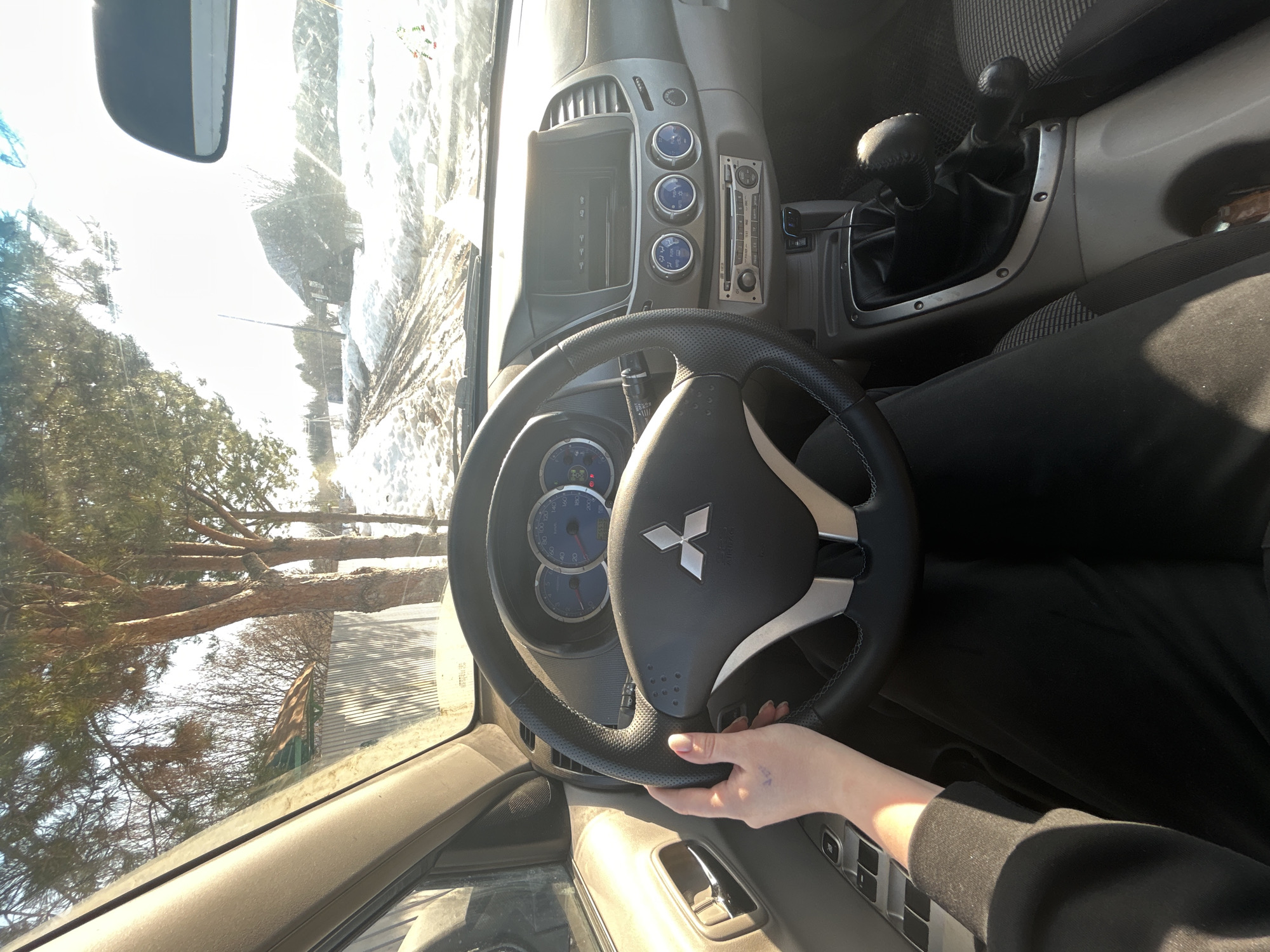 The image presents a first-person perspective from the driver's seat of a Mitsubishi vehicle, identified as being in the city of Фоки, Russia. The driver's right hand, with manicured nails, rests on the black leather steering wheel featuring the silver Mitsubishi emblem. The driver is wearing dark clothing, likely a long-sleeved top and pants.
The vehicle's interior shows a dashboard with blue-lit analog gauges (speedometer, tachometer) to the left, and a center console featuring a monochromatic display screen, three circular climate control knobs with blue illumination, and a car radio with a CD slot below. A manual gear shifter and a separate transfer case shifter are visible on the center console to the right of the driver's leg. Power window controls are present on the driver's door panel.
Through the windshield, a rural or semi-rural winter landscape is visible under bright sunlight. The unpaved road ahead is muddy with tire tracks and patches of melting snow. Snow covers the ground on both sides of the road. To the left, several pine trees and a corrugated metal fence are visible, with hints of a building behind them. To the right, another weathered or dark-colored building with a distinct roofline stands amidst the snow and sparse trees. Utility poles line the roadside.