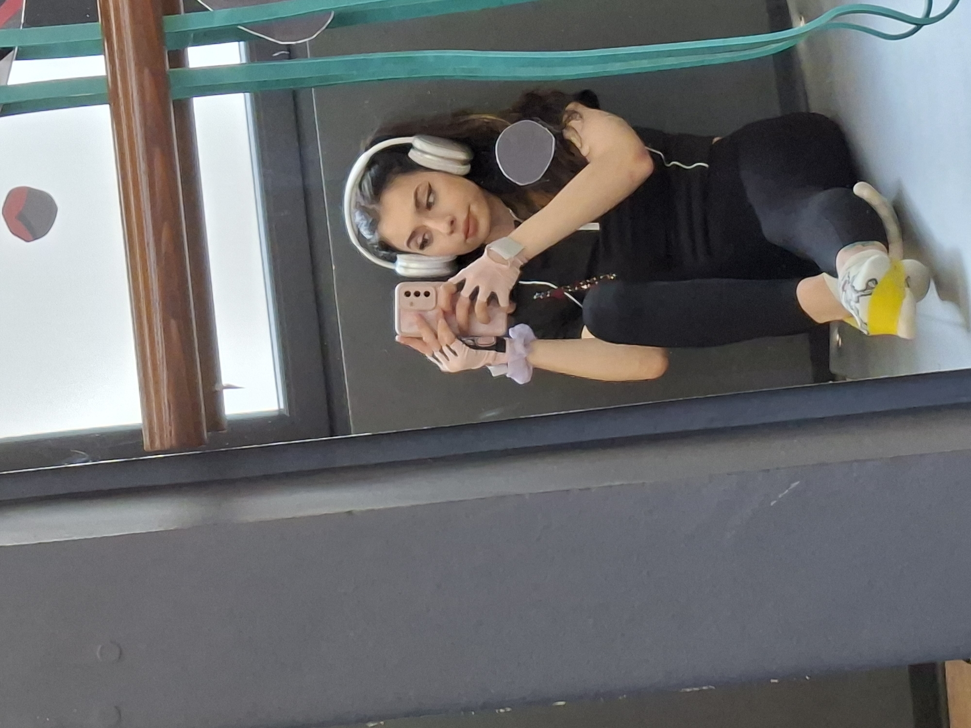An indoor, mirror selfie captures a young woman lying on her side, reflected in a dark, possibly polished floor or large mirror surface. She has long, wavy dark hair and is wearing white over-ear headphones, a black long-sleeved top, and black leggings. Her expression is neutral, with her gaze directed towards the smartphone she holds.
Both hands are adorned with light-colored fingerless gloves. Her left hand (on the right in the reflection) holds a light pink smartphone, which she is using to take the picture. A light-colored watch or fitness tracker is visible on her right wrist (left in the reflection). A light purple scrunchie is also on her left wrist. Two grey circular censor patches are digitally applied to the image, covering her chest area and upper thigh/hip area.
In the background, a teal or greenish-blue elastic band, possibly a resistance band, stretches horizontally. A tall, vertical wooden beam or pillar is visible on the left side of the reflection. Through what appears to be a window or glass partition in the upper left, a dark grey object with a red accent can be seen. The reflected environment includes a dark grey surface in the foreground and a lighter grey surface beyond, possibly a wall. There is no visible text in the image. The setting appears to be a modern indoor space, possibly a gym or a home with minimalist decor.