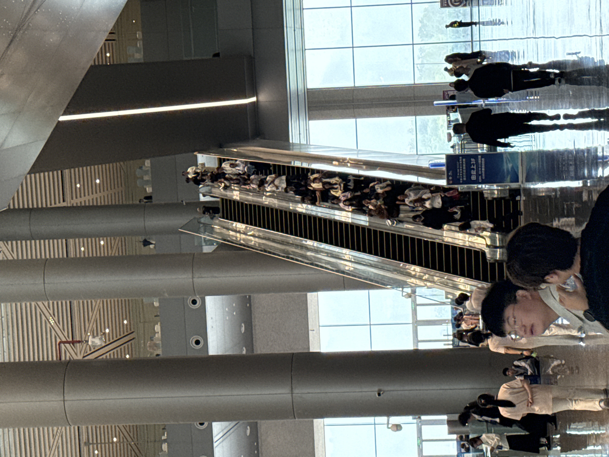The image shows an indoor public space with a large escalator. Many people are ascending the escalator. In the foreground, several individuals are walking or standing on the shiny, reflective floor. The environment appears to be a modern building, possibly a convention center or transportation hub, with tall columns and large windows in the background. The lighting is bright and diffused. The scene appears to be in the Chongqing High-tech Zone, China, given the context of the image's origin.