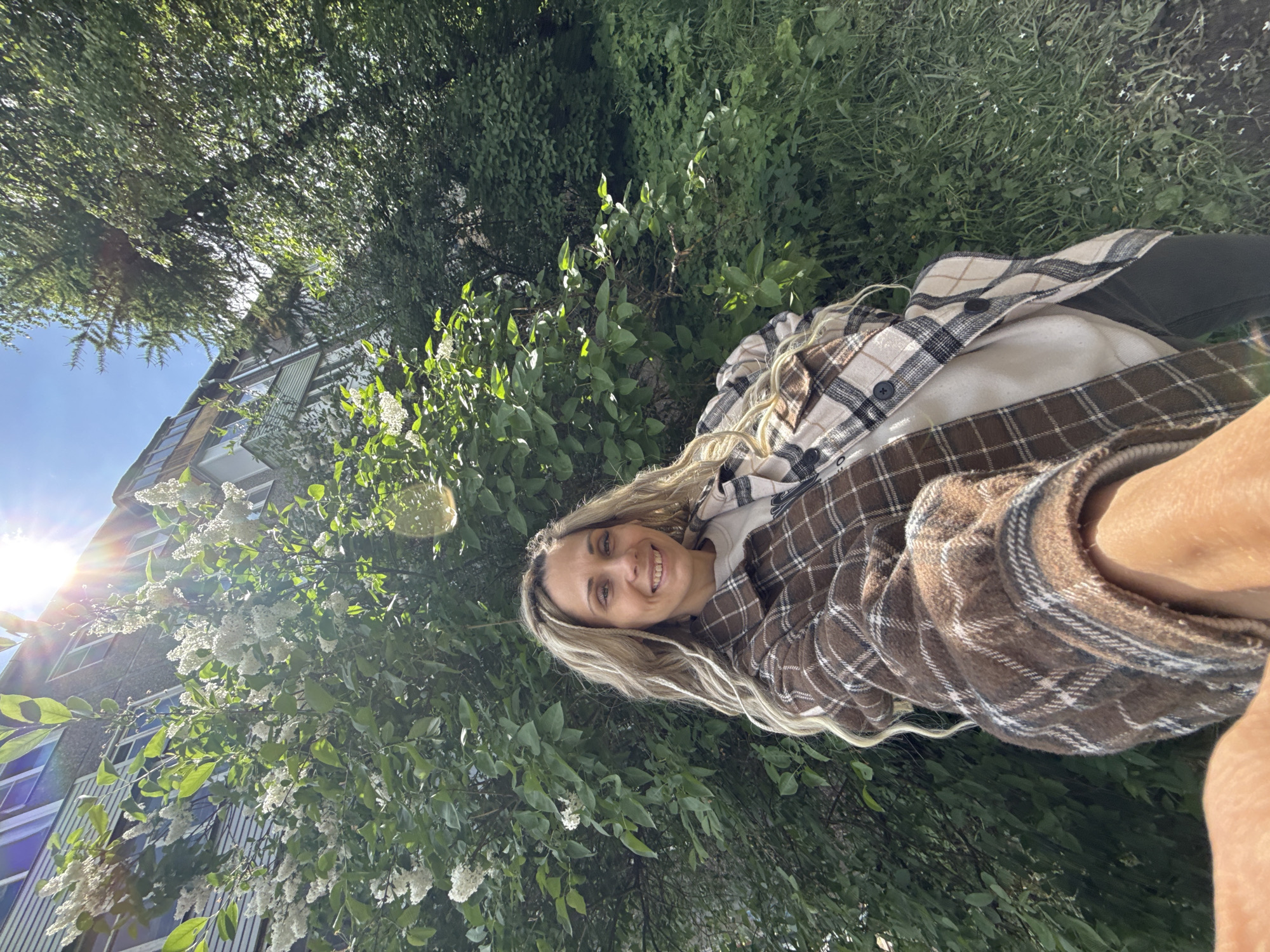 A woman with long, blonde hair is lying in the grass in a park. She is wearing a brown and white plaid shirt. The park is located in the city of Rusanovo, Russia. The surrounding environment includes trees, green grass, and a residential building. The subject appears to be taking a selfie, as her arm and hand are visible in the foreground.