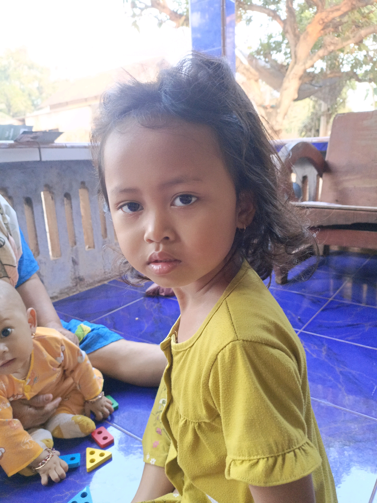 The image depicts two young children in Jenu, Indonesia. A girl with dark, wavy hair is in the foreground, wearing a yellow dress. To her left, a baby in an orange outfit is seated on a blue tiled floor, surrounded by colorful geometric toys. An adult's legs are visible between the two children, indicating their presence and likely supervision. The background shows a porch-like area with a concrete wall and a wooden chair, suggesting an outdoor or semi-outdoor domestic setting. The overall lighting is bright, indicating daytime.