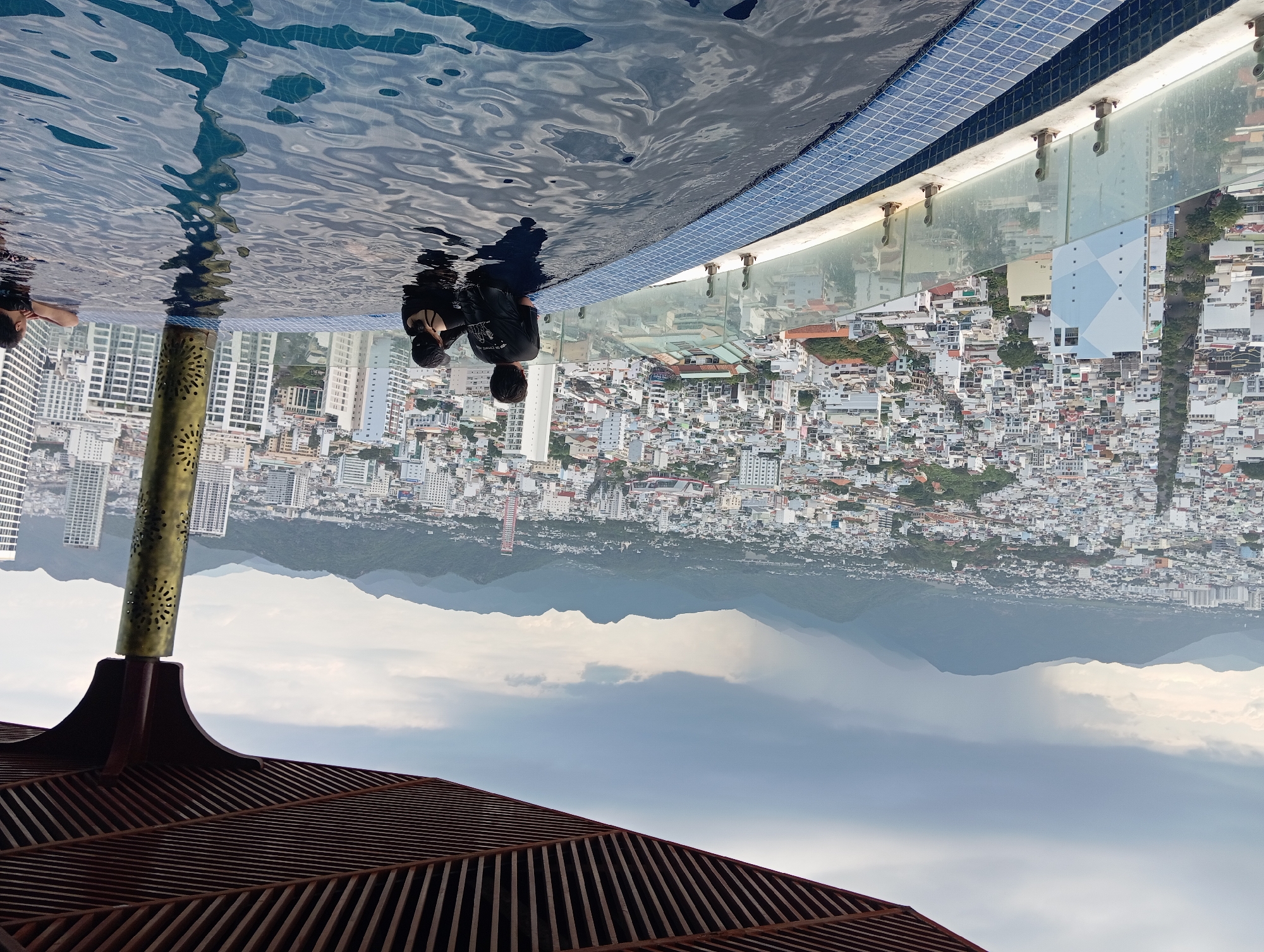 The image depicts an elevated swimming pool with an infinity edge, overlooking a dense cityscape and distant mountains under a cloudy sky. Two individuals, a man and a child, are partially submerged in the water, facing away from the camera. The man is wearing a black t-shirt, and the child is wearing a black shirt and a black cap. To the right, another person, whose face is not visible, rests their arms on the edge of the pool. The pool's edge is lined with blue mosaic tiles, and a glass railing with metal supports runs along the perimeter. The cityscape consists of numerous buildings of varying heights, some appearing to be residential, while others are taller, more modern structures. A brown, slatted overhang partially frames the top of the image. In the background, a range of green, mountainous terrain is visible, with a weather system suggesting potential rain or mist. The scene is suggestive of a rooftop pool in Nha Trang, Vietnam, likely offering panoramic views of the coastal city.