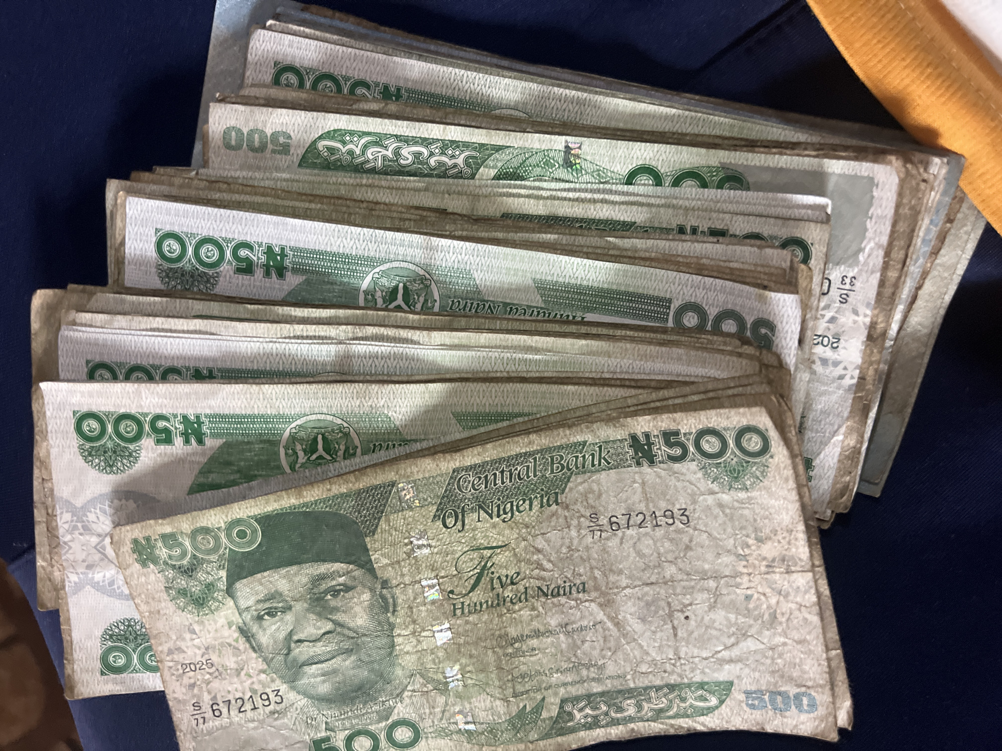 The image displays a stack of Nigerian 500 Naira banknotes, fanned out and overlapping. The banknotes are primarily green and white, with the denomination "500" clearly visible on many of them. The face of a man is prominent on the front of one of the visible banknotes, along with text that includes "Central Bank of Nigeria" and "Five Hundred Naira". The banknotes appear to be in used condition, with some creasing and signs of wear. The background is a dark blue fabric, possibly clothing or a surface. The lighting suggests an indoor setting, with some shadows cast on the banknotes. The image is a close-up shot, focusing on the details of the currency. The scene is static, with no apparent action or interaction occurring. The image does not contain any information that would suggest the location is in Nkpor, Nigeria.