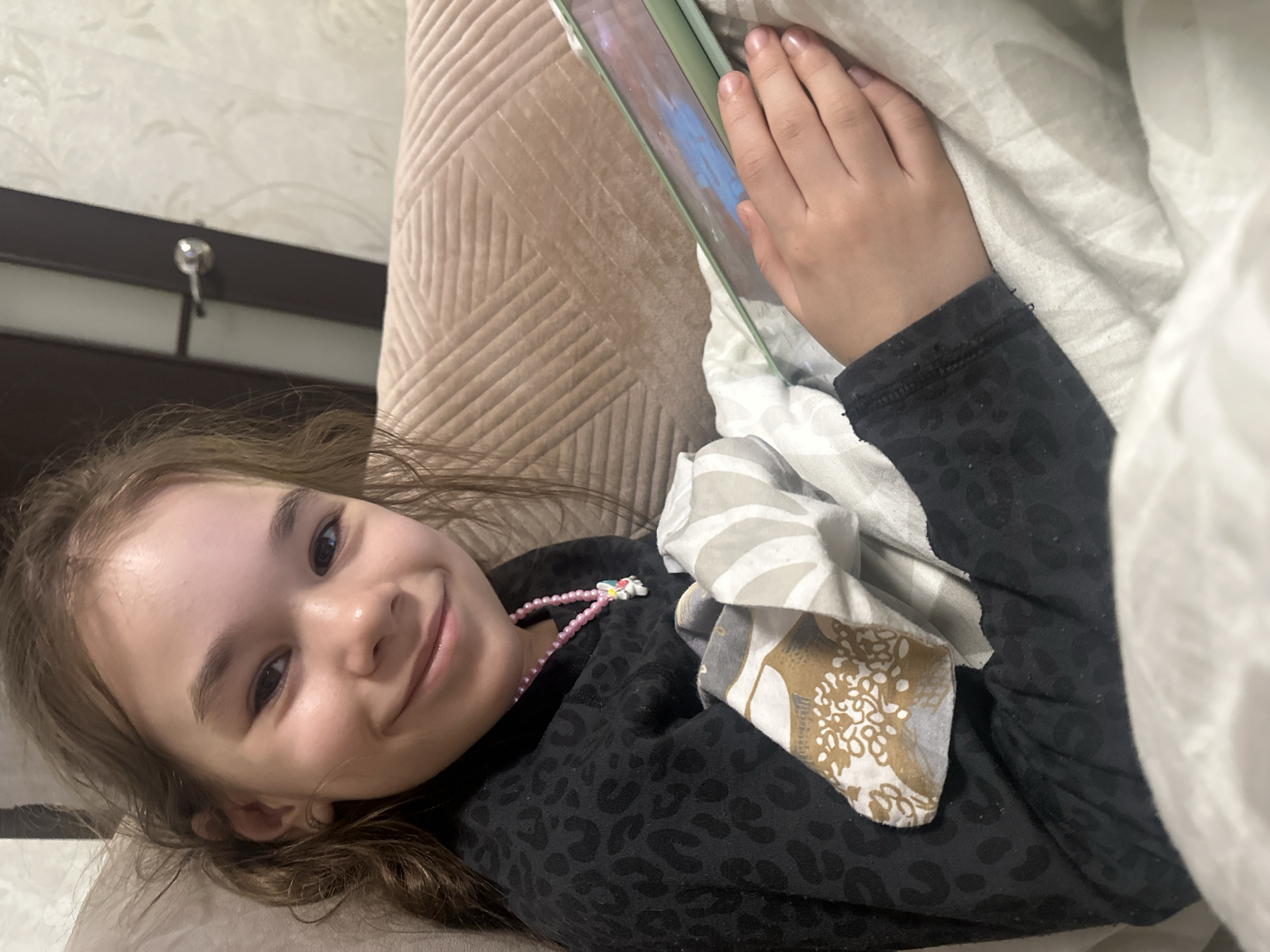 A young girl with light brown hair and a cheerful expression lies on her side, looking directly at the camera and smiling. She appears to be indoors, possibly on a bed or couch, with her head resting on a textured light brown surface.
She is wearing a dark, long-sleeved top with a subtle patterned design, resembling a leopard print. Around her neck is a pink beaded necklace with a small white charm, possibly an animal or cartoon character. Her right hand (from her perspective) is gently resting on a light green tablet or e-reader, which displays colorful content, predominantly blue and pink hues, on its screen. A white or light-colored blanket or duvet is partially covering her, with some patterned fabric visible near her left side.
The background shows a wall with a light-colored, subtle floral wallpaper pattern. Below the wallpaper, there's a dark horizontal structure, possibly a window frame or part of a furniture piece, from which a small metallic hook or latch protrudes. The lighting suggests an indoor setting, likely artificial, without clear indicators of time of day or weather beyond being inside. The overall mood is relaxed and content. There is no visible text in the image.
