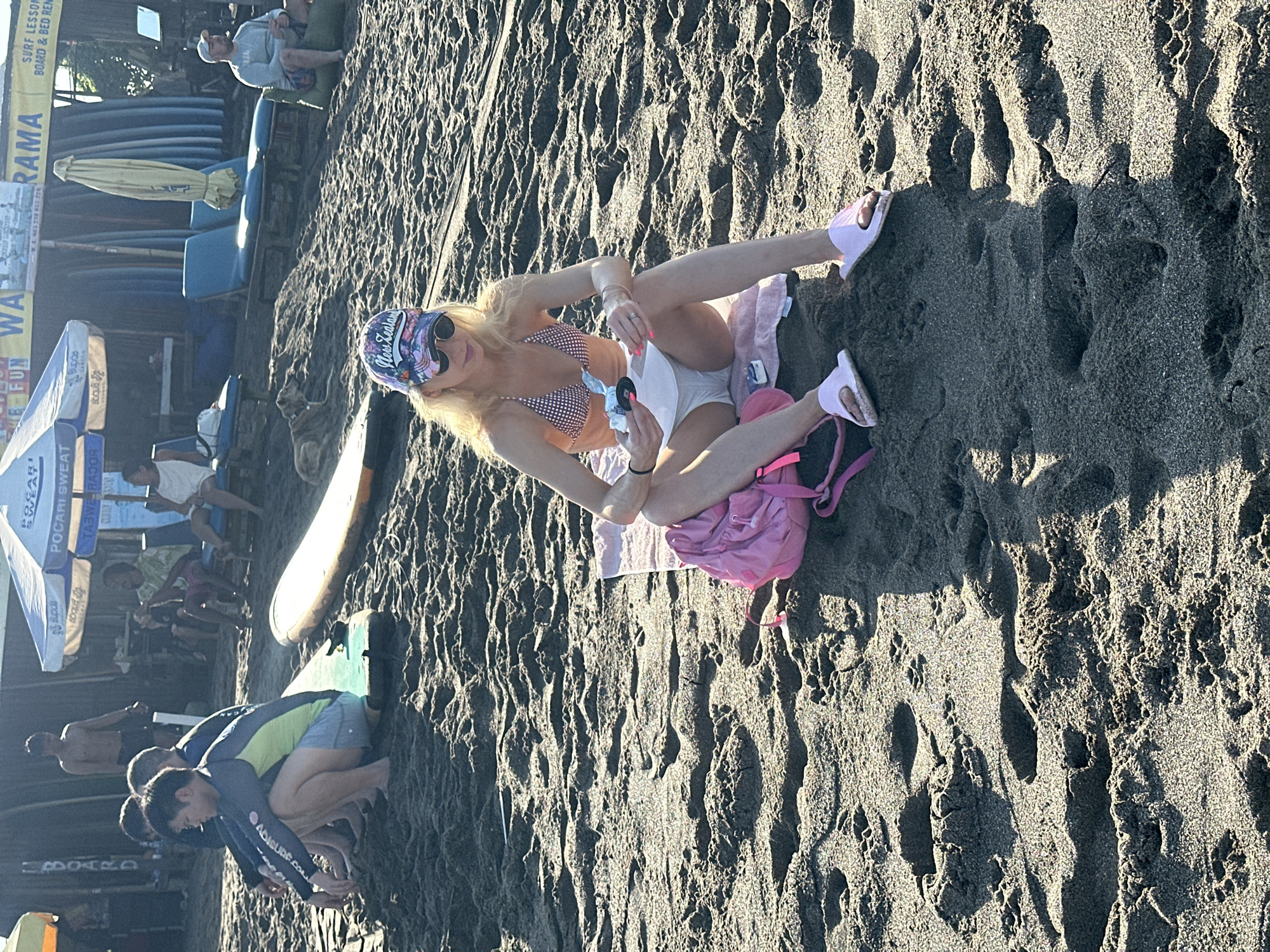 In North Kuta, Indonesia, on a beach with dark sand, a woman with blonde hair and sunglasses sits on a pink towel. She is wearing a checkered bikini top and a white skirt, and pink flip-flops. In her hands, she holds a small white object. A pink backpack rests beside her. In the background, several people are visible sitting on chairs and on the sand. Surfboards are lined up behind them, and beach umbrellas provide shade. The sun casts long shadows across the sand.