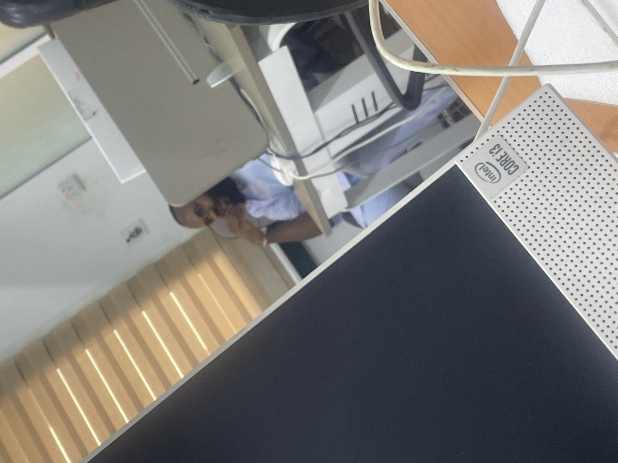 The image depicts an indoor scene, likely an office or study environment. A man wearing a light blue outfit is seated at a desk, looking towards a computer monitor. The monitor in the foreground, a white all-in-one desktop, displays an "Intel Core i3" sticker on its base. Behind the man, another computer monitor is visible, resting on a white desk. The background shows a wall with blinds casting striped shadows and a red indicator light. The overall impression is one of a functional workspace, possibly in an office building within Abuja, Nigeria, given the context provided.