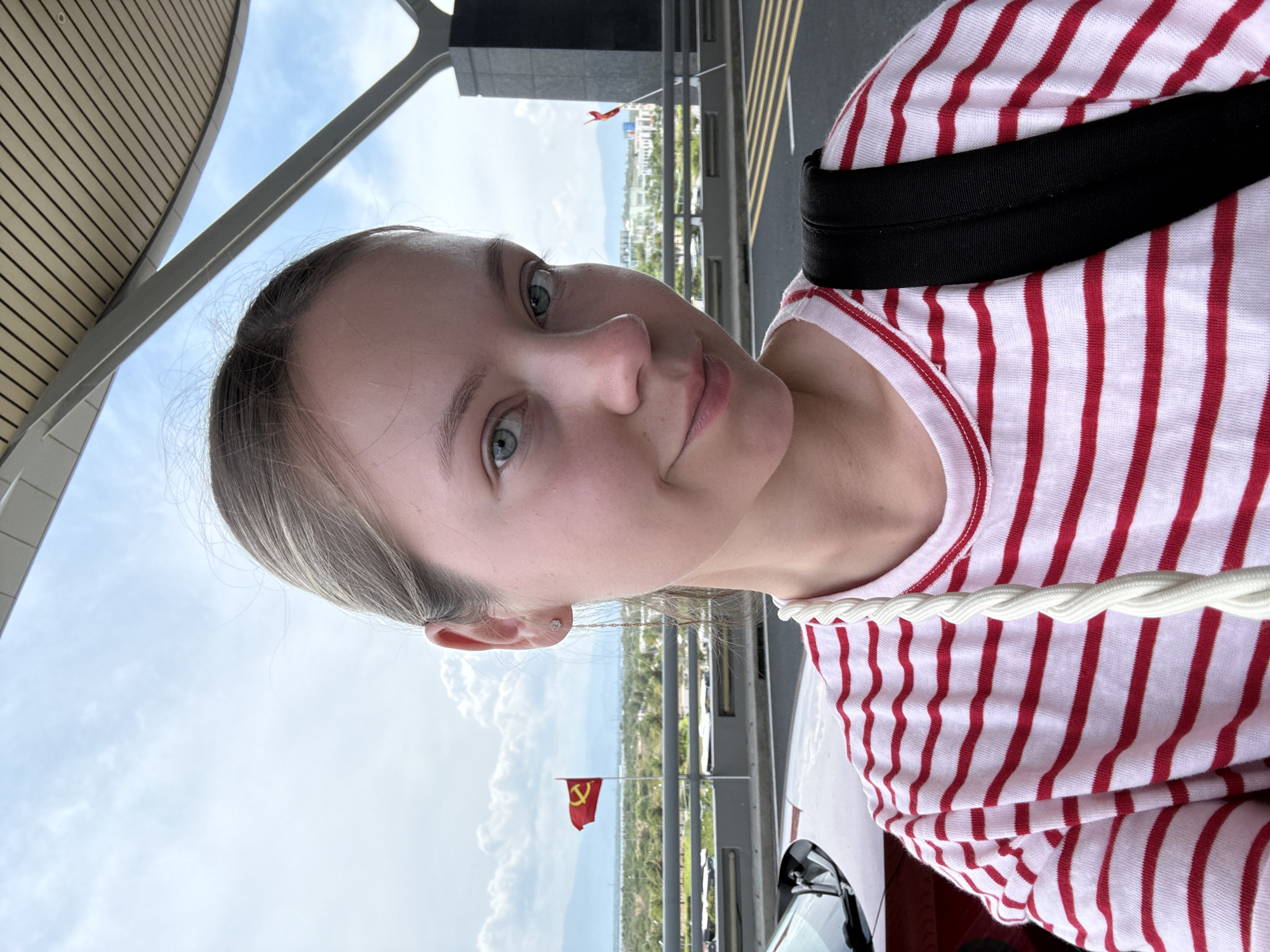 A medium shot depicts a fair-skinned woman in a red and white striped shirt with a black strap over her shoulder. She is standing outside, with a building structure above and a view of a cityscape in the background. A red flag with a hammer and sickle emblem is visible in the distance. The location is Bac Cam Ranh Ward, Vietnam, as indicated by the flag.