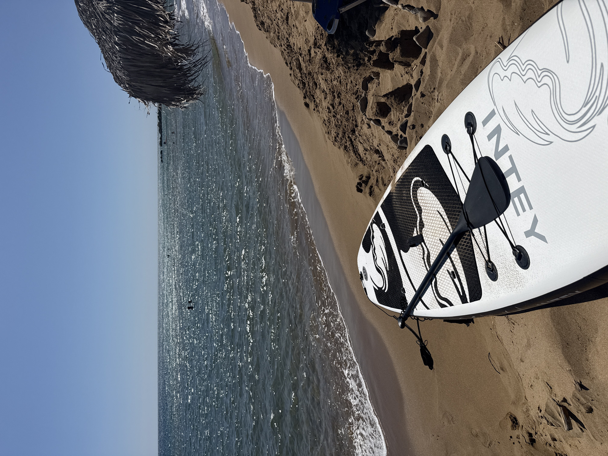 This is a photograph taken on a sunny day at a beach in the Municipal Unit of Lefkimmi, Greece. In the foreground, a white stand-up paddleboard with black graphics and the brand name "INTEY" is resting on the sand. A paddle is attached to the board with a coiled leash. The beach is sandy with some small pebbles and sandcastle-like structures. To the right, part of a thatched beach umbrella is visible. The sea is calm with gentle waves lapping at the shore. The water is sparkling under the sun, and several people are visible swimming or wading in the distance. The sky is clear and blue. The perspective is from a low angle, looking towards the sea.