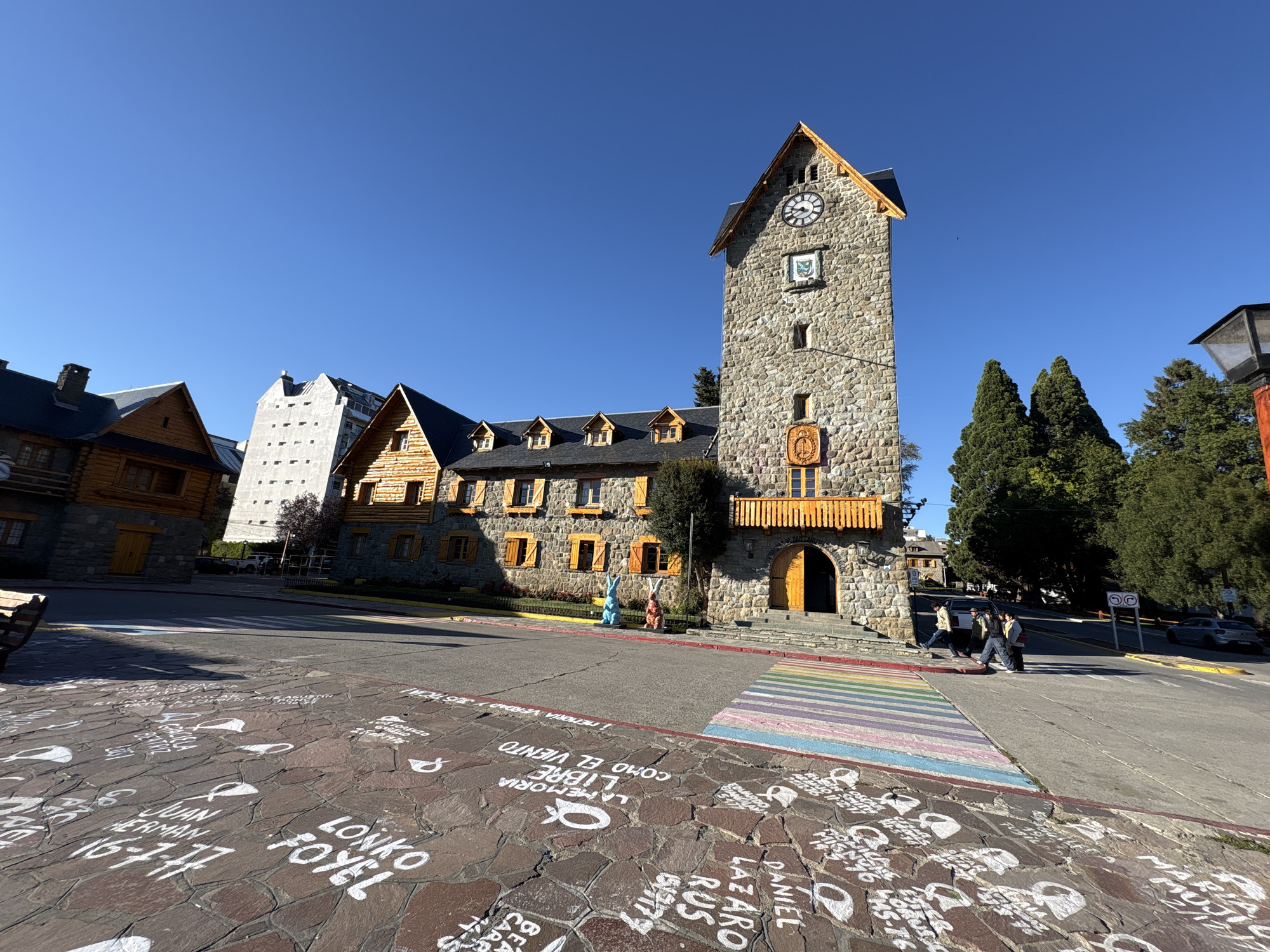 The image depicts a wide-angle view of the Civic Center in San Carlos de Bariloche, Argentina, under a clear, bright blue sky. The central subject is a multi-story stone building featuring a prominent clock tower with a round clock face, a wooden-shingled roof, and two crests positioned below the clock. The building's facade is constructed from irregular grey stone blocks, complemented by numerous rectangular windows with wooden frames and shutters. A large arched wooden entrance is visible at the base of the clock tower, flanked by a wooden balcony with vertical balustrades on the second floor.
In the immediate foreground, the ground is paved with reddish-brown irregular stones, covered extensively with white graffiti-style inscriptions and outlined figures. Readable text includes names and dates such as "LONKO FOYEL" and "JUAN HERMAN 16-7-77." A section of a crosswalk, painted with pastel horizontal stripes (pink, light blue, green, yellow, orange), crosses the asphalt street in front of the building.
On a grassy strip in front of the Civic Center's entrance, two large, stylized rabbit sculptures are positioned: one blue and one orange-brown. To the left of the main building, an adjacent log and stone structure with a dark roof and chimney is visible, followed by a taller, modern concrete building with square windows further back. To the far right, a dark lamppost with a square lantern top is partially visible.
Three individuals are observed walking across the street from right to left, away from the Civic Center. A dark SUV is parked on the far side of the street, with another vehicle further down. Tall, dark green coniferous trees are present behind the buildings and to the right, under direct sunlight that casts distinct shadows. The scene suggests daytime with optimal weather conditions.