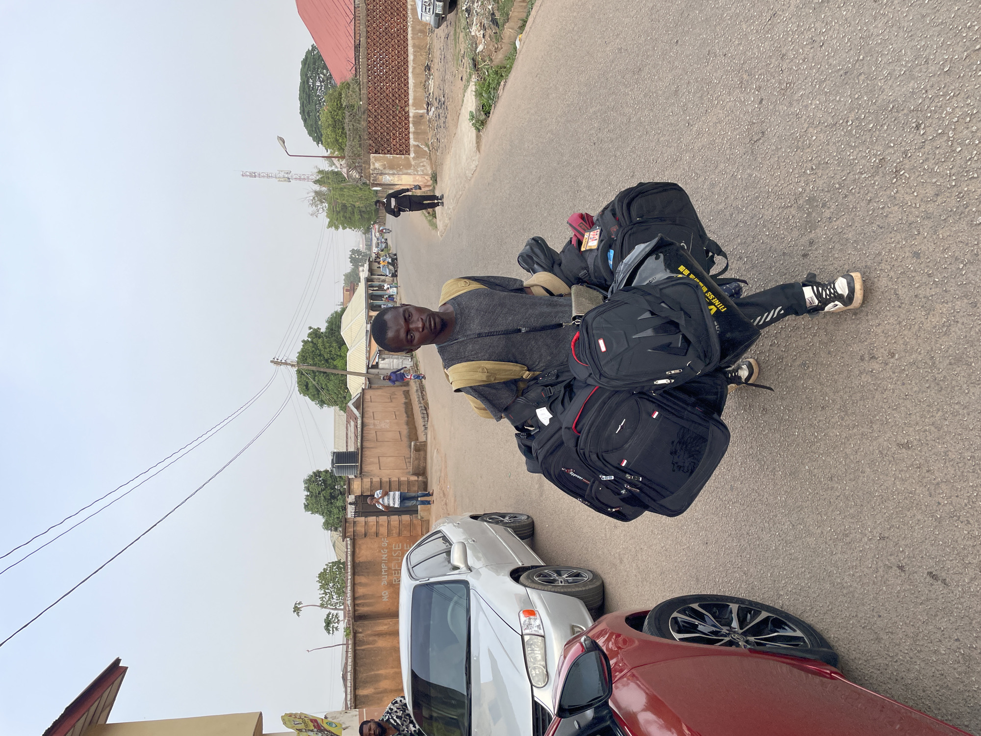 An individual stands on a paved street in Jos, Nigeria, surrounded by multiple black backpacks. The individual is facing the camera. They wear dark pants and a gray top with a tan vest. A silver car and a red car are parked at the side of the road. In the background, there are other people, buildings, and utility poles, indicating an urban environment. The sky is overcast.