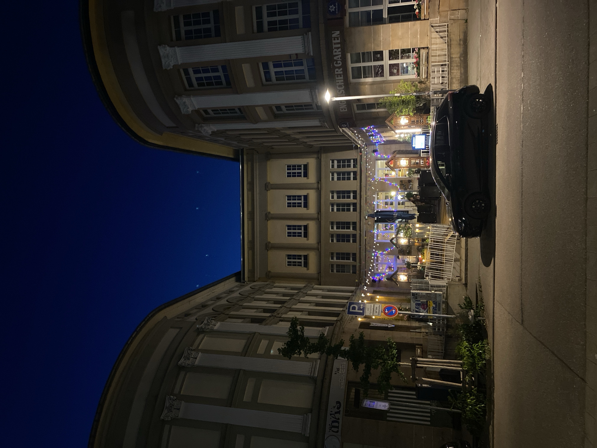 The image shows a street scene at night in Landau in der Pfalz, Germany. The view looks toward a restaurant area nestled between two curved buildings, their architecture featuring multiple windows and decorative columns. The sky above the buildings is a deep blue with a few visible stars. The restaurant area is illuminated with string lights, and tables and chairs are visible. A black car is parked along the right side of the street. Street signs are posted near the base of one of the buildings.