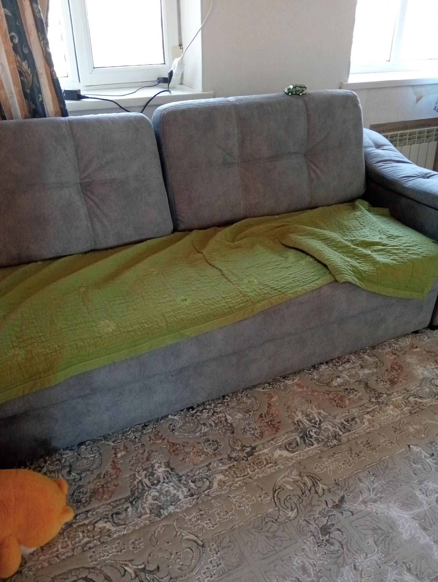 A plush gray sofa is positioned in front of a window with light-colored curtains. A bright green blanket is draped over the sofa. A small toy truck is visible on the back of the sofa. The floor is covered with a patterned rug. A portion of an orange stuffed animal is in the lower left corner of the image. There is a radiator visible to the right of the sofa. Electrical outlets and wires are visible on the windowsill.