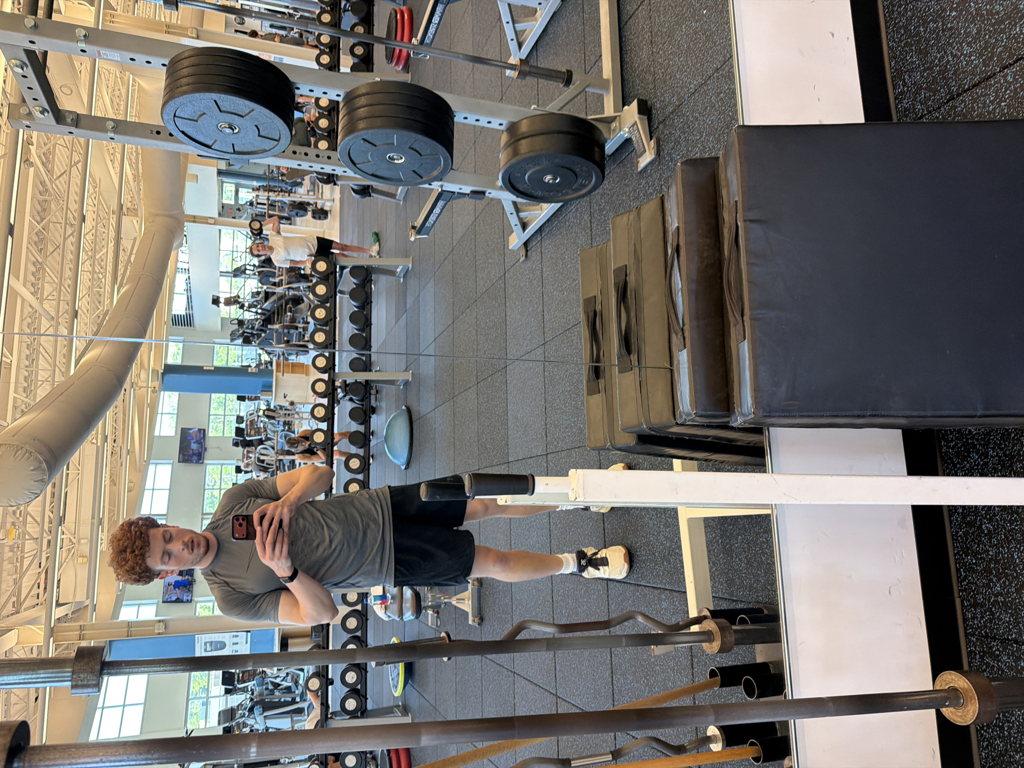 This is a full-body, mirror selfie taken by a young adult male with curly red hair, wearing a gray t-shirt, black athletic shorts, white socks, and light-colored athletic shoes. He is standing in a gym, holding a smartphone and looking down at it. The gym has a rubberized floor and racks of weight plates and barbells are visible. In the background, other gym-goers can be seen exercising, and exercise equipment like treadmills and weight machines are present. There are also large inflatable tubes on the ceiling. In the foreground, a white vertical support post is visible, and to the right, a stack of black exercise mats is positioned. A large mirror covers a significant portion of the left side of the frame, reflecting the gym environment and the person taking the selfie. The location appears to be a fitness facility in Beachwood, Ohio, United States.
