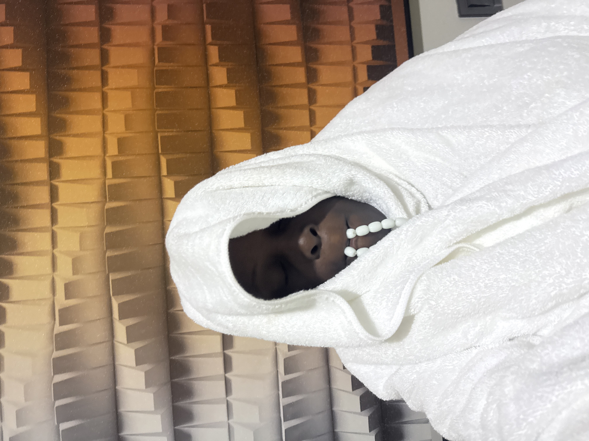 A person wearing a white bathrobe and head covering is shown with closed eyes and a string of white beads in their mouth. The background is a textured wall with a gradient of beige to orange colors. The scene is indoors.