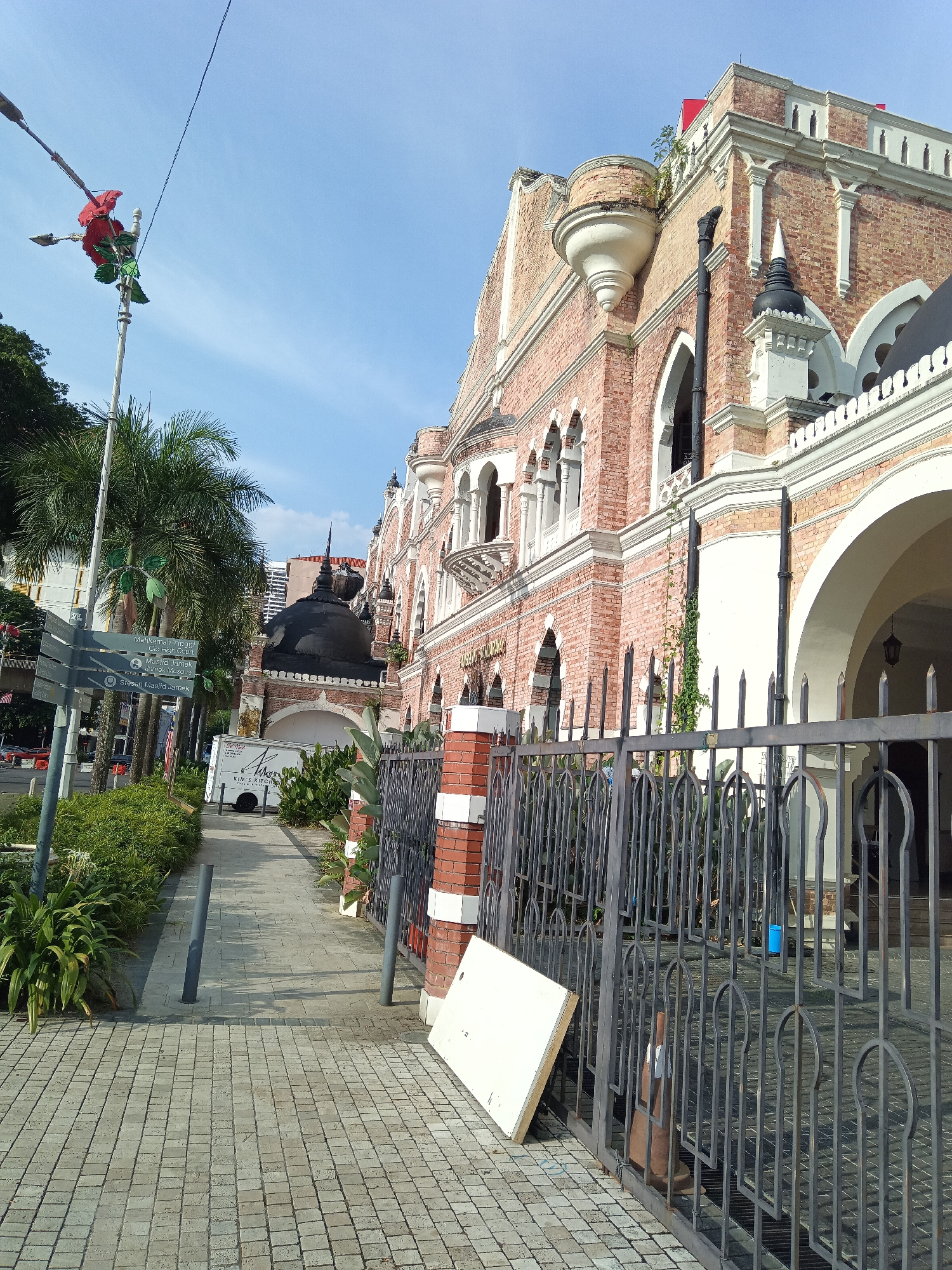 The image shows a view of the Sultan Abdul Samad Building in Kuala Lumpur, Malaysia. The building, constructed in red brick with white ornamentation, features multiple arches, balconies, and a prominent black dome. In the foreground, a metal fence with decorative arches runs along the sidewalk. A paved walkway leads past lush green plants and a directional signpost indicating "Mahkamah Tinggi" (High Court), "Masjid Jamek" (Jamek Mosque), and "Stesen Masjid Jamek" (Jamek Mosque Station). A white delivery truck with "KIM'S KITCHEN" printed on its side is parked on the sidewalk. Overhead, a street lamp with a red rose decoration is visible against a clear blue sky. The scene depicts a sunny day with clear skies.