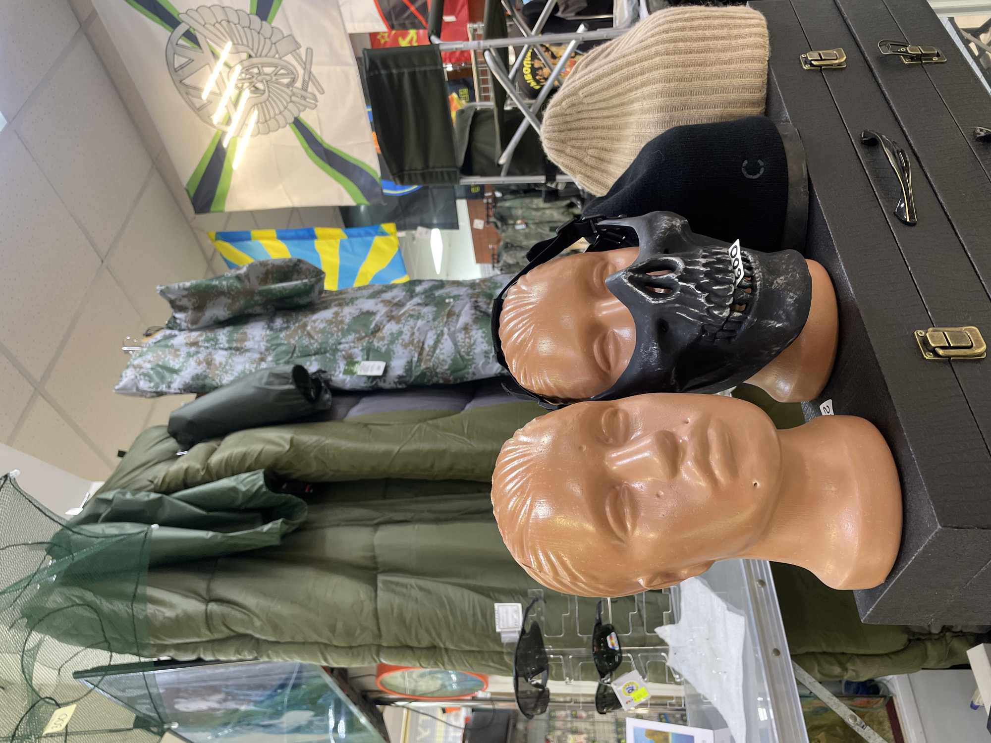 The image shows a retail display inside a store, likely specializing in military or outdoor equipment, potentially located in Krasnoyarsk, Russia, although this cannot be definitively confirmed from the visual data alone. The display includes:
- Two tan mannequin heads, one wearing a black skull mask and a black beanie.
- A beige knitted beanie.
- Several sleeping bags in green and camouflage patterns.
- A case with metallic latches.
- A flag or banner with a coat of arms.
- A fishing net.
Other items are visible in the background, including additional flags, bags, and assorted merchandise. The overall environment suggests a retail setting.