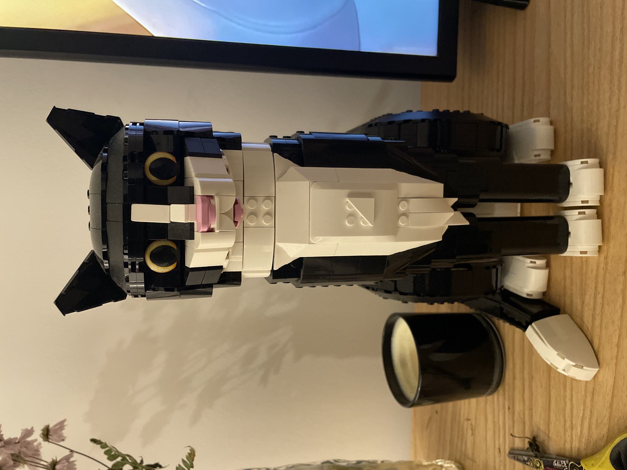 The image shows a Lego cat model, primarily black and white, on a wooden surface. The cat is lying down, with its head towards the top left of the frame. A black cylindrical object, possibly a candle, is next to the cat model on the right side. The cat's details include pink nose and golden eyes. In the background, part of a television screen is visible above the Lego model. A portion of foliage, possibly flowers, appears at the bottom left of the image. A pair of scissors are visible on the bottom right. There is no indication of the location being Buenos Aires, Argentina.