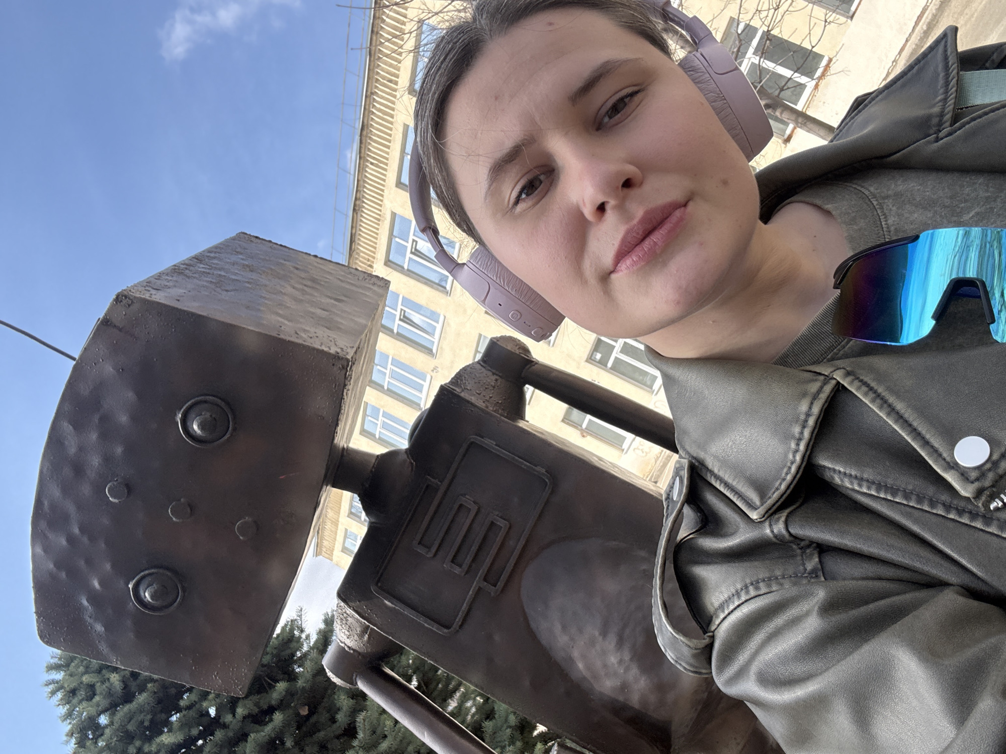 A young woman poses for a selfie with a metal sculpture of a robot in Chaikovsky, Russia. The robot has a square head with two round eyes and a rectangular body. The woman wears headphones and a leather jacket. The background includes a building with windows and a tree. The sky is blue with some clouds. The woman is on the right side of the image, and the robot sculpture is on the left. The camera angle is low, looking up. Sunglasses are hanging on the woman's jacket.