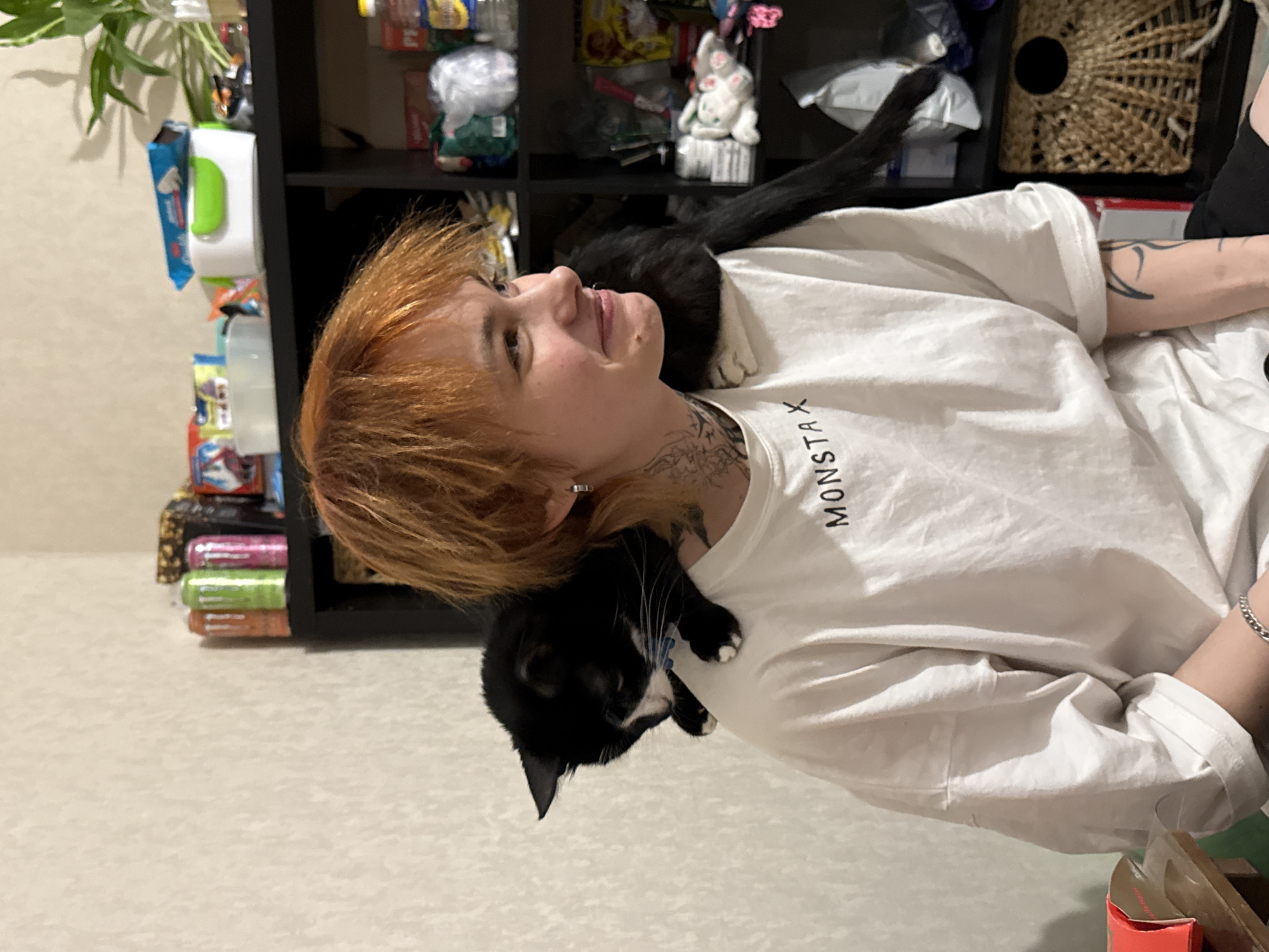 An indoor residential scene in Saint Petersburg, Russia, depicts a young individual and a cat. The person, positioned slightly left of center and facing right in profile, has short, shaggy, reddish-orange hair and is smiling, revealing a dimple on their right cheek. They wear a white, loose-fitting t-shirt with "MONSTAX" printed in black capital letters across the chest. A silver hoop earring is visible in their left ear, along with a small piercing on their right nostril. Tattoos are present on their neck, extending downwards from behind the left ear, and on their left forearm. A silver chain bracelet adorns their left wrist.
A black and white tuxedo cat with white paws and a visible light blue collar is perched on the individual's left shoulder, with its head lowered, looking towards the individual's chest. Its tail extends down the person's back.
In the background, a dark, multi-shelved unit, likely a bookcase, holds various items. The top shelf displays several colorful beverage cans (orange, green, pink), a gold-toned decorative item, a white rectangular container with a green lid, and a green potted plant. The middle shelf contains assorted plastic containers, packages, and bottles, including a blue and white package atop the white container. The bottom shelf features a woven wicker basket with a circular opening, alongside other miscellaneous objects. The wall behind the individual is light-colored and textured. A wooden surface and a red object are partially visible in the lower-left foreground.
