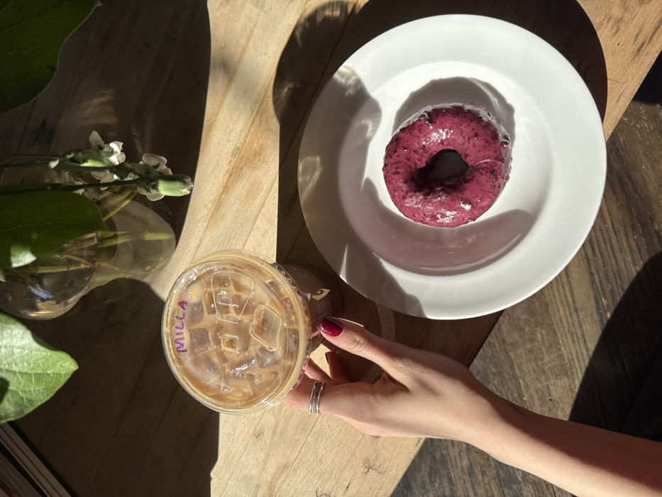 An overhead view captures a person's left hand interacting with a beverage and food item on a light wooden table. The hand, featuring dark red manicured nails and two rings on the fourth finger, holds a clear plastic cup filled with a light brown, milky iced beverage and ice cubes. The name "MILLA" is written in purple marker on the cup's clear plastic dome lid. To the right, a single circular donut, covered in a glossy deep purple, speckled glaze, rests on a plain white ceramic plate. In the upper left background, a section of a clear glass vase containing green foliage and slender stems with small white flowers is visible. Strong natural light illuminates the scene, casting distinct shadows across the wooden surface. The environment is an indoor establishment, likely a cafe, located in New York, United States of America. The precise address or name of the establishment cannot be determined from the visual information presented.