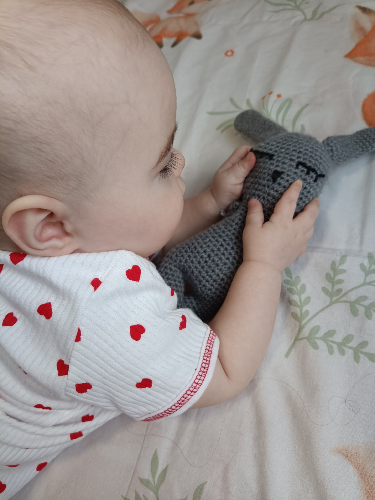 An overhead shot captures a baby lying down, gently holding a grey crochet bunny toy. The baby is seen from the side, with their head, ear, and hands in clear view. They have fair skin, fine light hair, and noticeable long eyelashes. The baby is wearing a white, ribbed long-sleeved top adorned with numerous small red heart patterns. Their small hands are clasped around the soft, grey, crocheted bunny, which has simple black stitched eyes and a nose, giving it a peaceful, sleepy appearance.
The background is a light-colored blanket or sheet patterned with whimsical cartoon foxes in shades of orange and green leafy sprigs, suggesting a playful or nursery setting. The lighting is bright and even, indicating either daytime or a well-lit indoor environment. A few loose strands of hair are visible on the blanket near the baby's arm. The baby appears calm and engaged with the toy. No visible text is present in the image. The location context for this scene is Noviy, Russia.