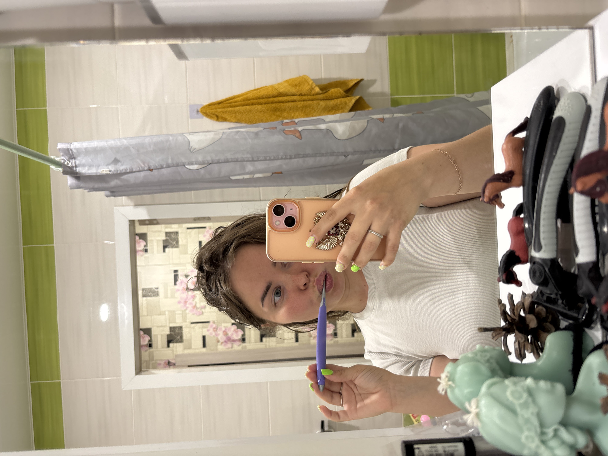 A mirror selfie is captured in what appears to be a bathroom, reflecting a person holding a purple dental tool to their lips and a peach-colored iPhone to take the photo. The person has light skin and shoulder-length brown hair and is wearing a white t-shirt. The bathroom features horizontally stacked light-colored tiles with vertical green stripes, a grey shower curtain with a faint pattern, and a mustard yellow towel. In the lower right corner, various hair accessories and decorative items are visible on a white surface. A framed wall decoration of a light-colored pattern with pink floral accents is visible in the reflection of the mirror. The location is most likely in Izhevsk, Russia.