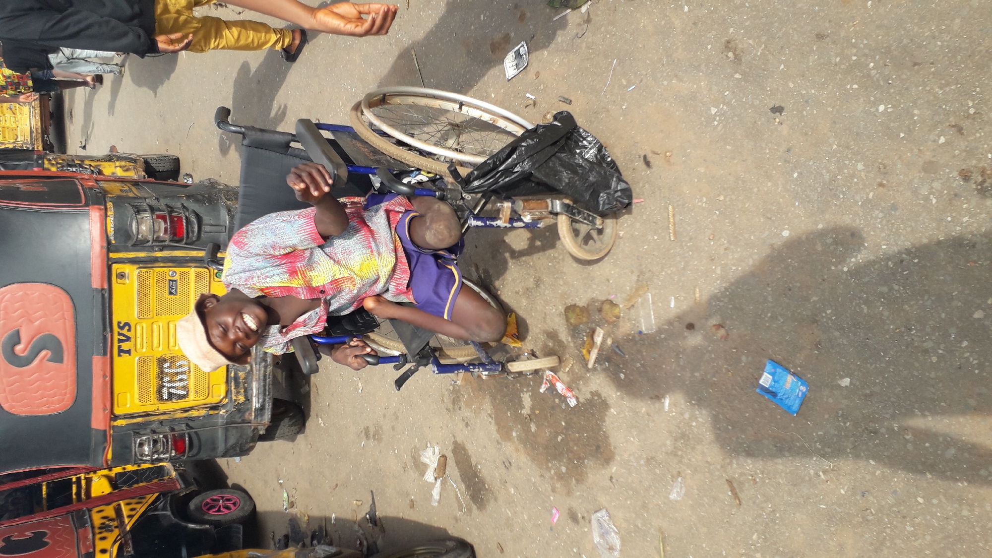 This media file captures a scene in Nkpor, Nigeria, possibly during the daytime under fair weather conditions. The primary subject is a young man in a wheelchair, sitting beside a yellow and black motorized tricycle (also known as an auto-rickshaw or "keke"). The man is wearing a colorful, patterned short-sleeved shirt, purple shorts, and a light-colored cap. He is smiling broadly and looking towards the camera, appearing cheerful and engaged. His wheelchair is blue and appears to be a standard model, with a black plastic bag placed on its seat.
Behind the man and his wheelchair is the rear of a TVS brand auto-rickshaw. The auto-rickshaw is predominantly yellow with black accents and has a large red "S" logo on its side panel. Visible text on the auto-rickshaw includes "TVS" and a license plate that reads "TRC 786". Another similar auto-rickshaw is partially visible in the foreground on the left.
The setting appears to be a street or roadside, with a concrete or asphalt surface that shows signs of wear and scattered debris, including plastic wrappers and other small items. The overall impression is a snapshot of daily life in a bustling Nigerian locality, with the man in the wheelchair being the central focus, conveying a sense of joy and resilience.