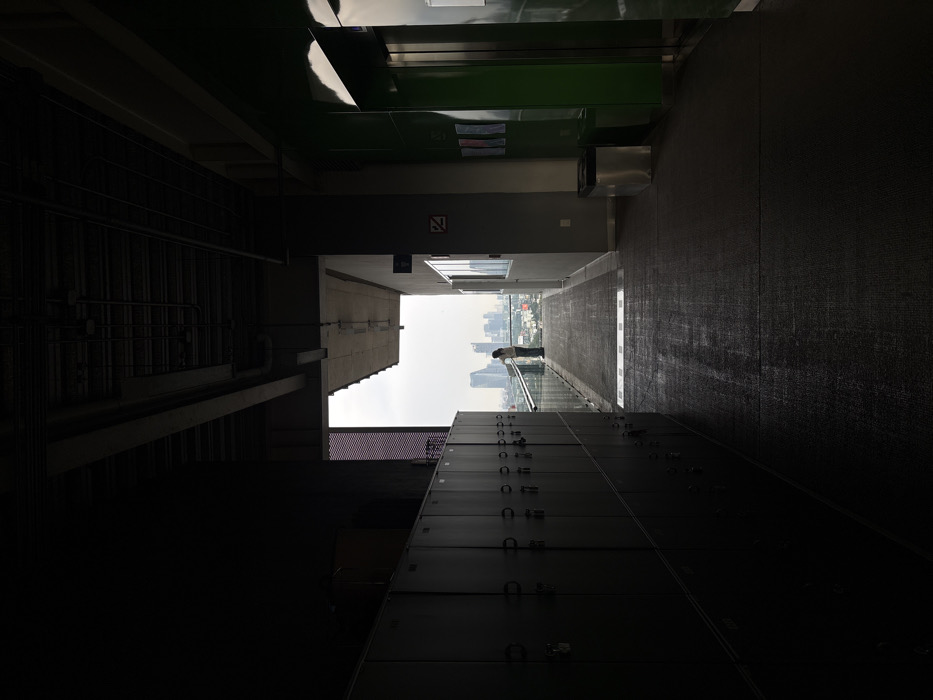 The image displays an interior hallway transitioning to an outdoor balcony overlooking a cityscape in Mexico City, Mexico. On the left, a long row of dark grey metal lockers, arranged in two horizontal tiers, extends from the foreground. The upper wall and ceiling on the left are dark, revealing exposed pipes and structural elements. On the right, a grey wall segment features a white window panel and a red warning sign. Further right are two bright green vertical panels, likely elevator doors, adjacent to a silver cylindrical trash receptacle. The ceiling on this side also shows visible infrastructure. The dark, textured floor leads towards the brightly lit exterior. At the far end of the balcony, a single person, dressed in light clothing, stands facing away from the camera, leaning on a clear glass railing. This individual is silhouetted against an overcast sky. Beyond the railing, a dense urban skyline composed of numerous high-rise buildings extends into the distance under a hazy sky.