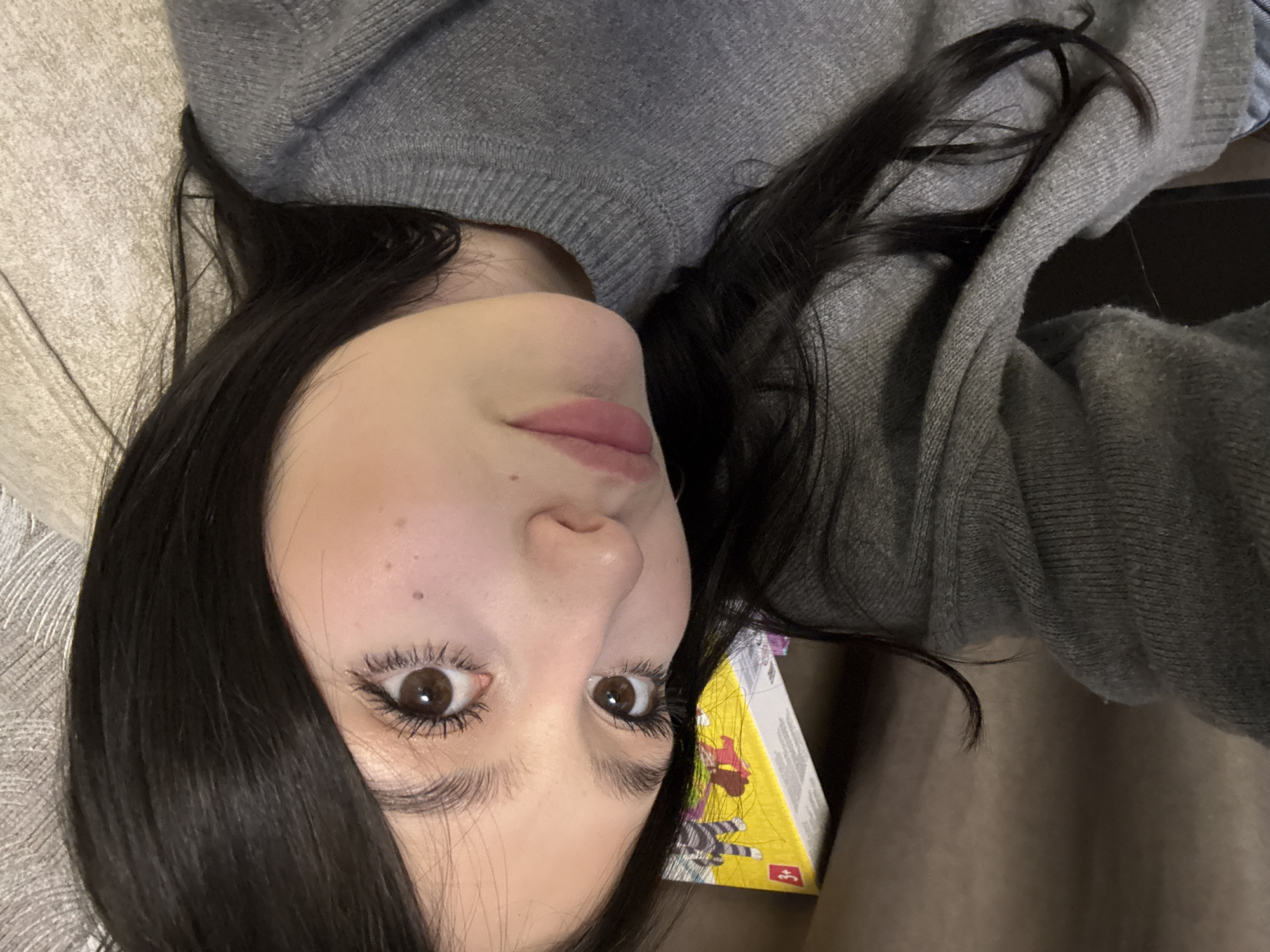 A young female, appearing to be in an indoor residential setting, is depicted in a close-up, head-and-shoulders portrait. She has long, dark hair, dark brown eyes, and light skin with a faint mole or freckle below her right eye. Her eyelashes appear enhanced, and her lips are a light pink hue, slightly parted. She is wearing a grey knitted sweater. The individual is positioned slightly angled, looking directly towards the camera, indicative of a selfie. The background to her left features a dark brown, textured surface, possibly a cushion or sofa arm. To her right, a lighter grey, textured surface is visible, also suggesting soft furnishings. Partially visible behind her upper right head and shoulder is a yellow box with white edges, displaying a graphic that includes a striped figure and what appears to be the number "3". There are no visual cues within the image to identify the specific city as Фоки, Russia, or any other geographical location.