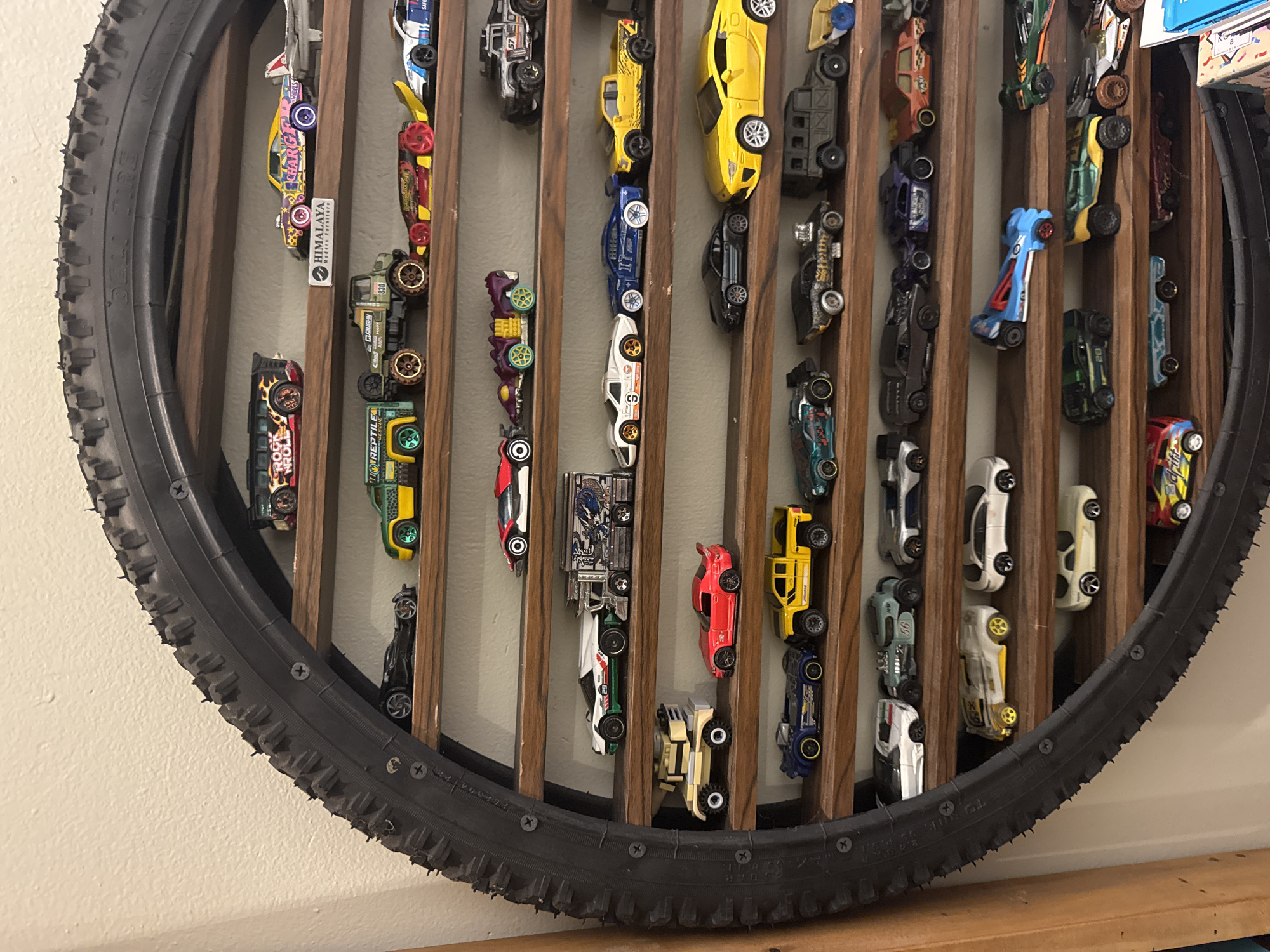 A circular tire frame, mounted on a wall and resting on a wooden surface, houses an array of vertically arranged toy cars. The tire, textured with treads, is affixed to a wooden structure with vertical slats, creating compartments for each toy car. The toy cars vary in color and design, including a yellow sports car, a blue race car, a red truck, and several others with various markings and styles. The background is a plain, light-colored wall. Text labels, such as "HIMALAYA" and "CHARGE," are visible on some of the cars. The overall composition suggests a display or collection of toy cars within a DIY frame. There is no indication of the city of Kuta Selatan, Indonesia, in the provided information.