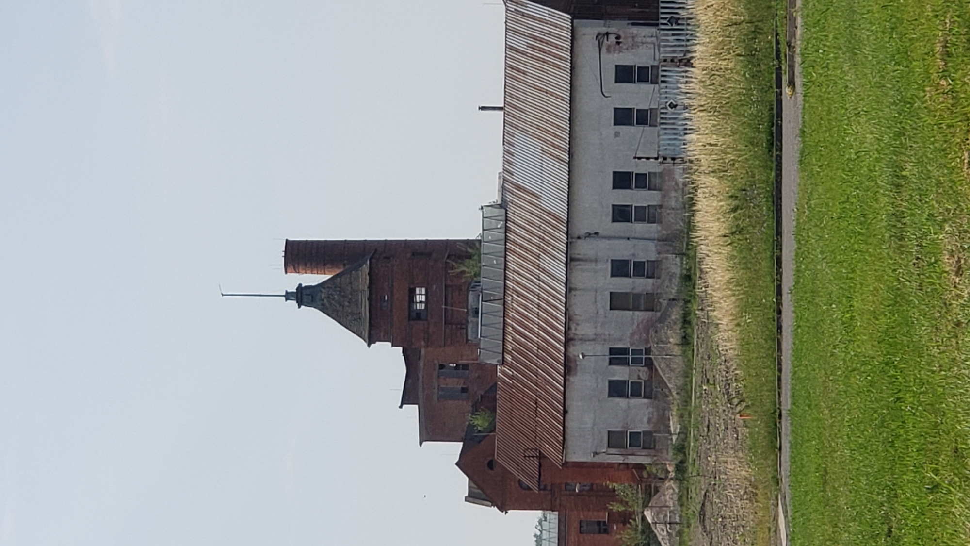The image shows a cluster of industrial brick buildings under a clear sky. The main structure on the right features a tall, cylindrical brick chimney and a smaller, ornate tower with a pointed roof. To the left of this, several other brick structures are visible, some with broken windows and overgrowth. In the foreground, a long, rectangular building with a weathered, corrugated metal roof runs horizontally across the frame. This building has numerous small, evenly spaced windows. The ground in front of this building is covered with dry, tall grass and some patchy bare earth. A section of manicured green lawn is in the very front of the image. The overall impression is of an abandoned or disused industrial site. This industrial complex is located in the Solivars district of Prešov, Slovakia, historically known for its salt mining and processing.
