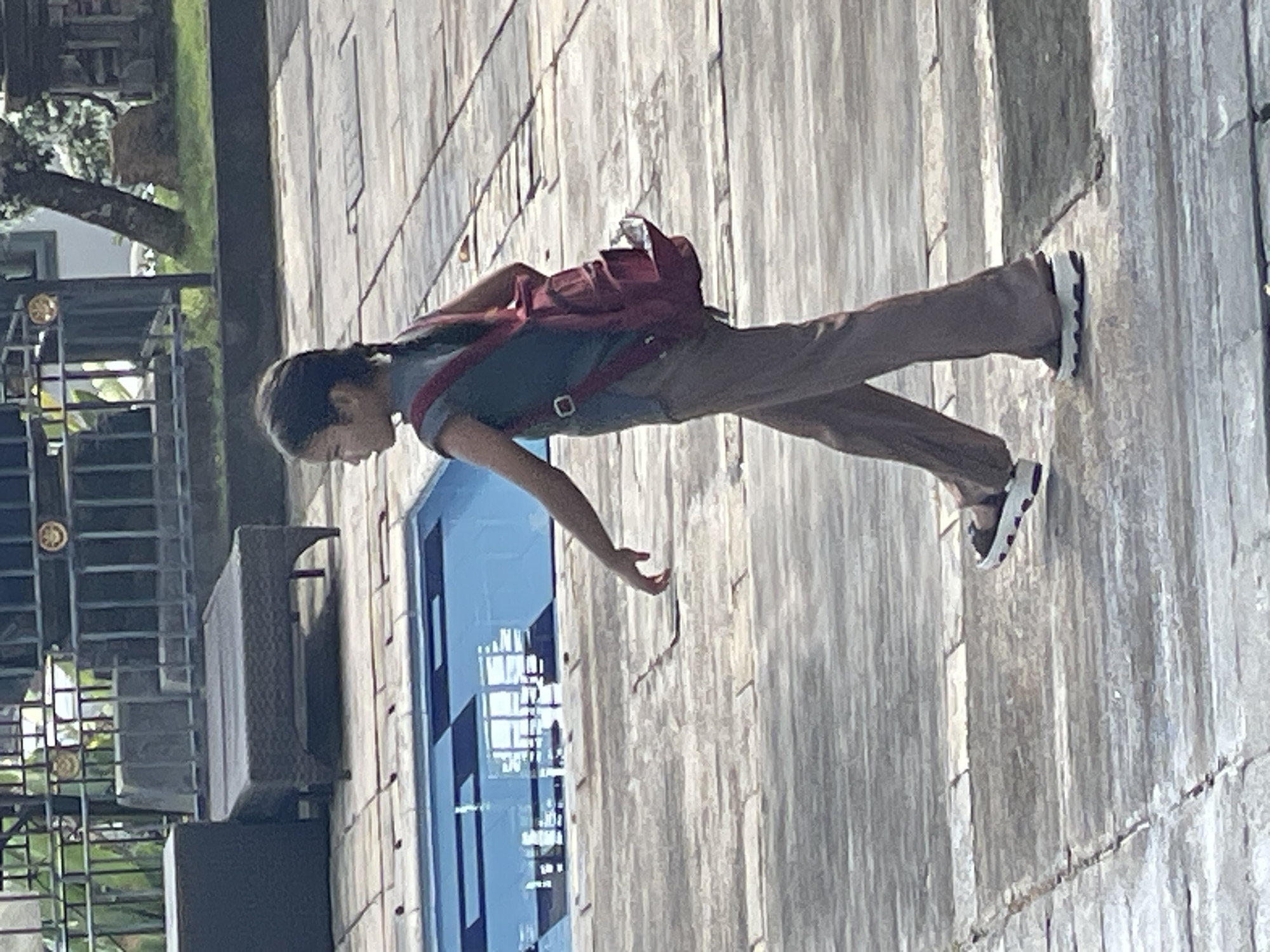 A young girl with dark hair in a braid walks across a tiled patio, her body angled to the right. She wears a sleeveless dark blue top, brown bell-bottom pants, and white platform sandals. A red backpack with a silver water bottle sticking out of it is slung over her left shoulder. In the background, a portion of a swimming pool with blue tiles is visible, along with outdoor furniture and a dark metal fence. The setting appears to be Kuta Selatan, Indonesia.