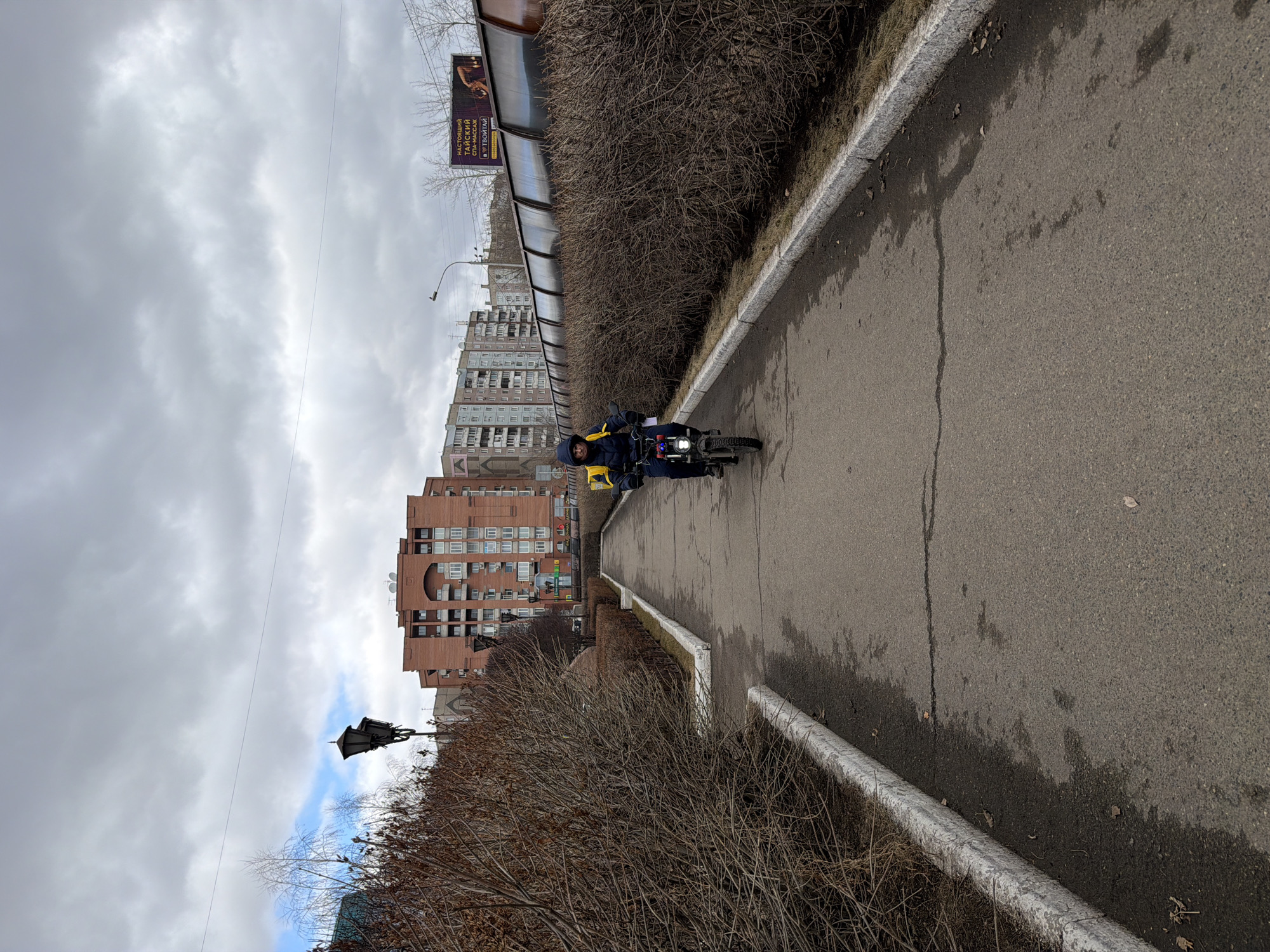 An urban scene depicts a person riding a black motor-scooter or moped on an asphalt path. The person is wearing a dark jacket or hoodie, dark pants, and has a bright yellow bag or backpack on their back. The scooter has a prominent headlight and some red and blue details on its front.
The paved path, showing some wear and cracks, runs alongside a low, white concrete curb that separates it from a dense line of leafless, brown bushes, suggesting late winter or early spring. Beyond the bushes, a translucent barrier or fence is visible. In the background, several multi-story residential buildings with brick facades and numerous windows rise under an overcast sky. One building features a distinctive arched architectural element. A tall streetlight stands to the left of the path.
The sky is predominantly cloudy with patches of brighter, whiter clouds, and a hint of blue sky is visible in one section. There are wet patches on the asphalt, indicating recent precipitation or melting.
A billboard is visible in the upper right part of the image, displaying an image that appears to be of an exotic animal (possibly a scorpion or similar) and Russian text. The text reads: "НАСТОЯЩИЙ ТАЙСКИЙ СПА-МАССАЖ В ТВОЙТАЙ," which translates to "REAL THAI SPA-MASSAGE IN TVOYTAY." The setting appears to be a typical urban environment in a city like Krasnoyarsk, Russia.