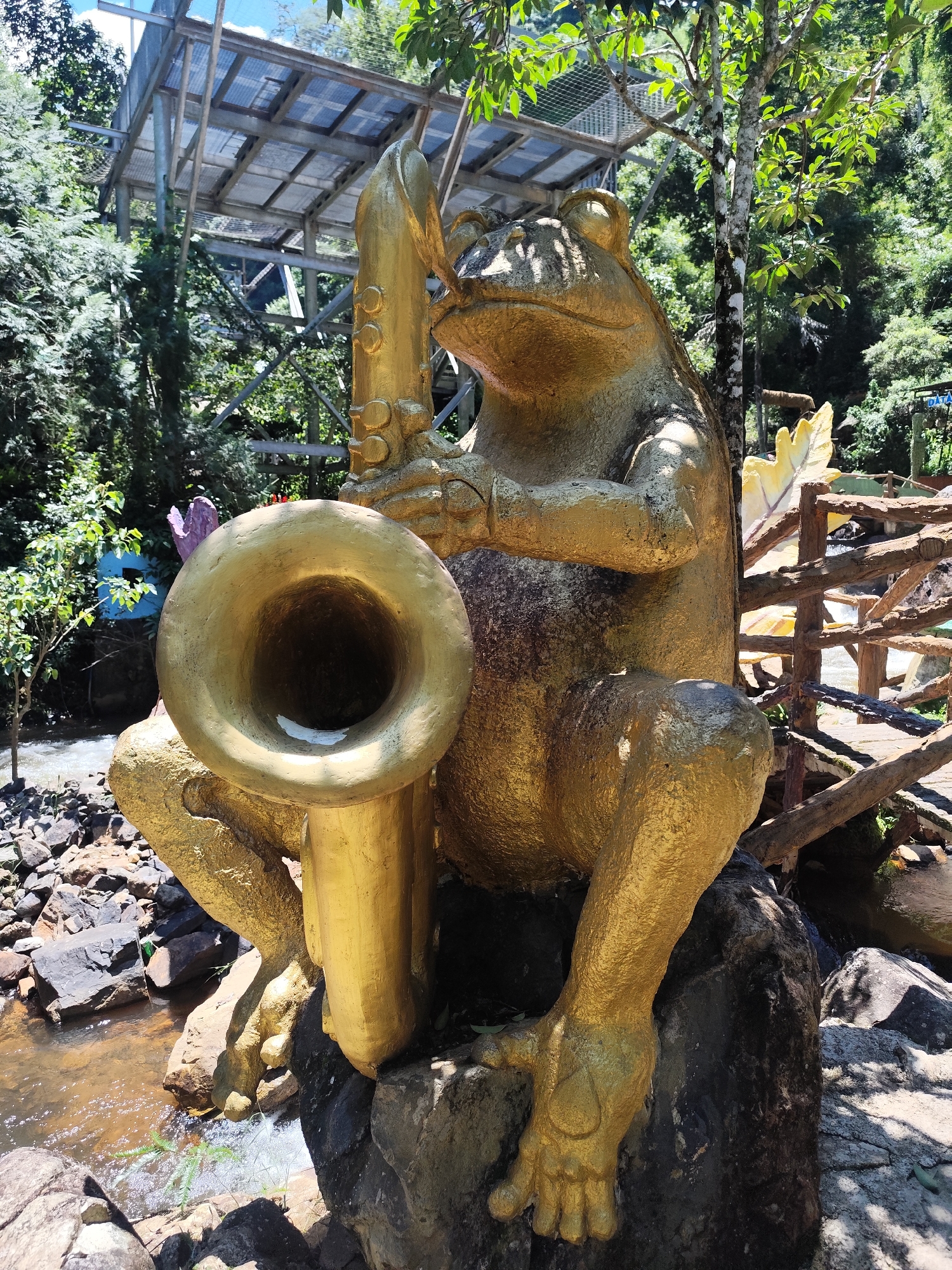A life-size, golden statue of a frog playing a saxophone sits on rocks near a stream in Da Lat, Vietnam. The frog is depicted in a seated position, holding the instrument with its front legs. The statue is weathered and has visible textures, suggesting it is made of a durable material. Behind the statue, lush green trees and foliage dominate the background, with a rustic wooden fence and a raised walkway structure visible in the mid-ground. Sunlight filters through the trees, casting shadows on the statue and the surrounding rocks. The stream flows with clear water over smooth stones. The scene suggests an outdoor tourist attraction or park environment.