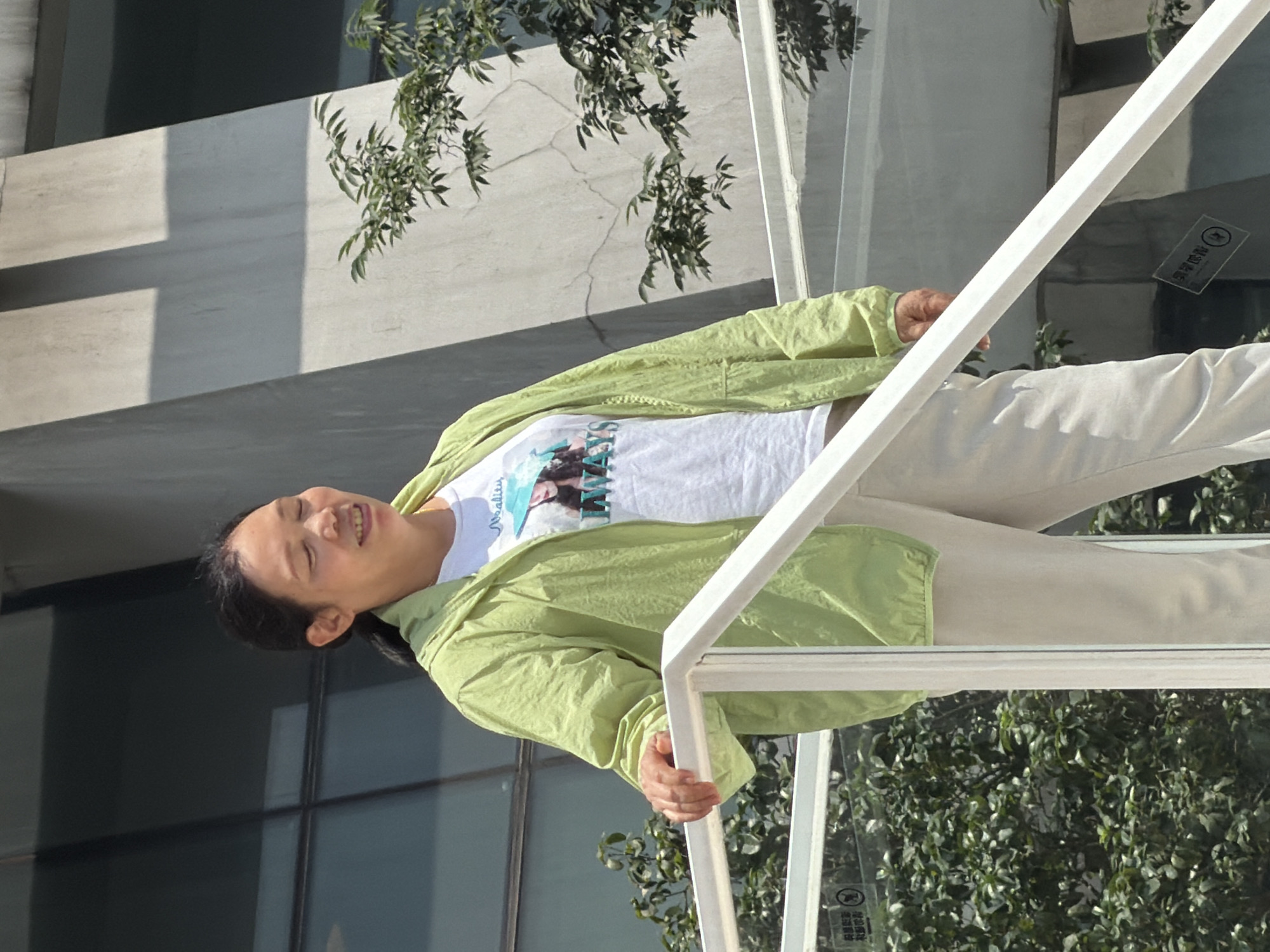 An East Asian woman with dark hair pulled back in a ponytail stands on a white railing on an outdoor staircase, wearing a light green hooded jacket over a white t-shirt with a graphic print and light-colored pants. Her eyes are closed and she appears to be smiling. Green foliage is visible behind her, and the background includes a modern building with geometric architectural elements and large windows. The scene appears to be in the Yuzhong District, China, indicated by signage in Chinese on the railing.
